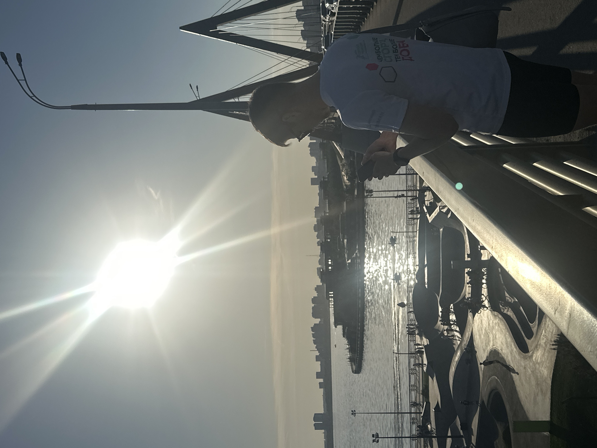 The image shows a person standing on the Millennium Bridge in Kazan, Russia, looking over the railing towards the Kazanka River. The sun is brightly shining in the sky, creating a glare on the water. The Kazan skyline is visible in the background, but somewhat obscured by the bright light. The person is wearing a white t-shirt with what appears to be Russian text and a logo. They are also wearing shorts. The bridge structure, including its cables and architectural details, are visible. The person is holding a phone or device in their hands and looking at it.