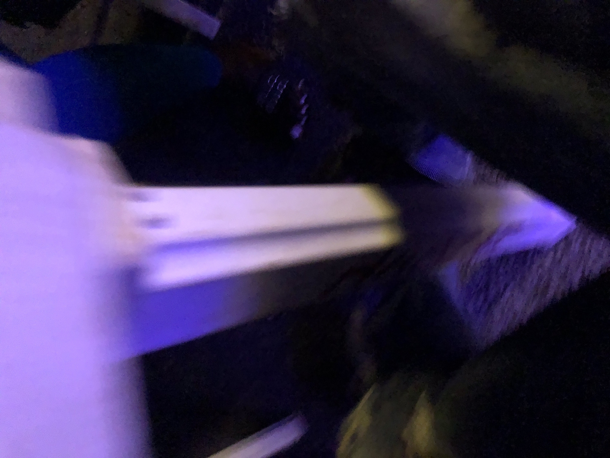 An extreme close-up, low-angle, and vertically oriented photograph taken in the city of Unknown, Unknown, depicts a distorted and abstract scene dominated by blurred shapes and vibrant purple and blue lighting. The primary subject appears to be a white, vertical structure, possibly a pole or a pillar, that recedes into the background. The structure is illuminated from above with a strong purple light, casting a luminous glow and creating soft shadows. To the right of the structure, a dark, possibly blue, object is visible, also blurred. The foreground is obscured by a dark, blurry mass, suggesting the presence of the photographer's legs or knees, or perhaps a dark surface. The background is indistinct and appears to be a mix of dark textures and subtle, scattered lights. The overall impression is one of movement and abstract forms due to the motion blur and intense lighting.