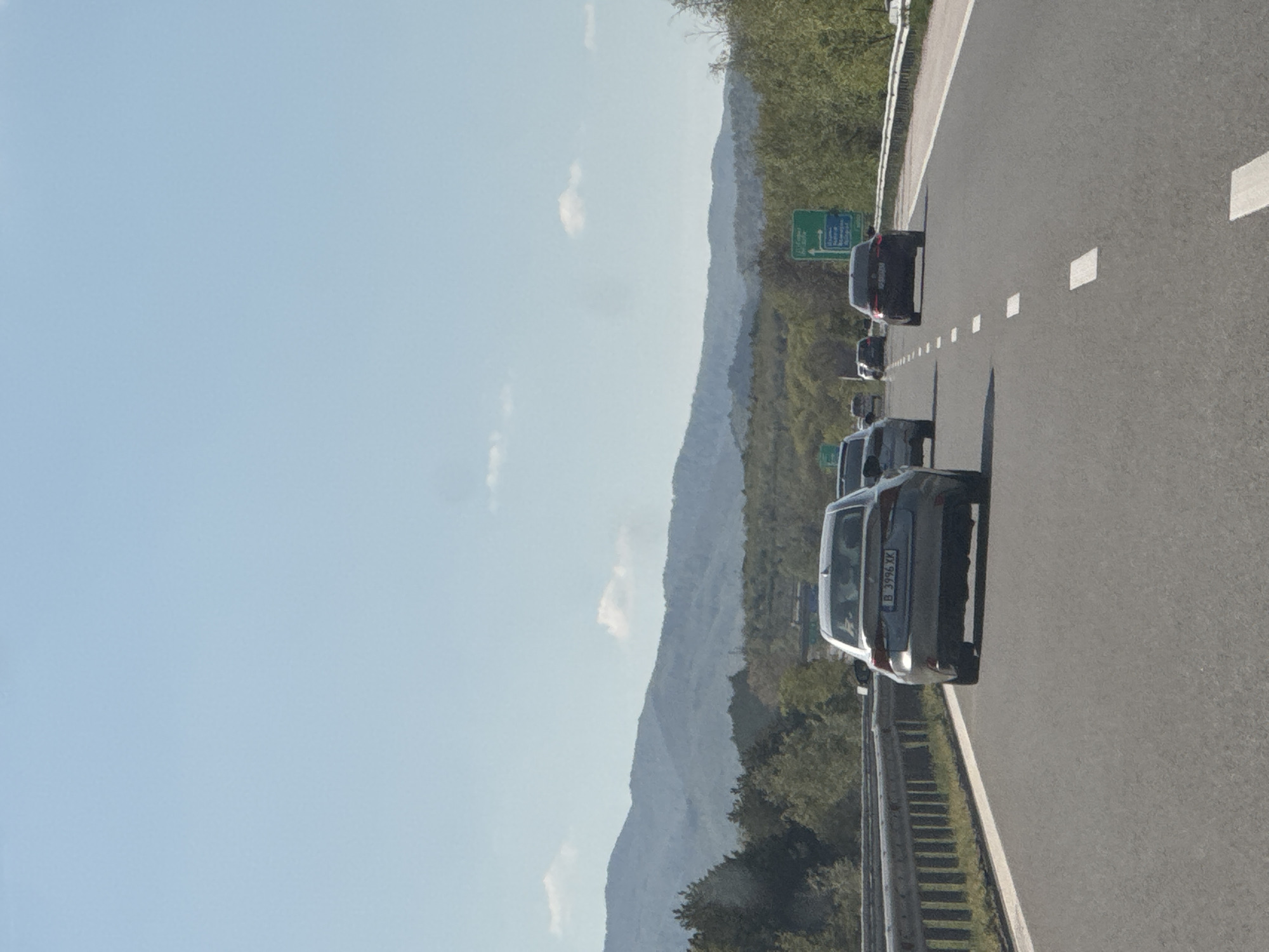 The image, correctly oriented, displays a daytime scene on a highway near Lesichovo, Bulgaria. The sky is bright and mostly clear with a few wispy clouds. The highway itself is an asphalt road with prominent white lane markings, extending into the distance.
Several cars are visible on the highway, forming a queue or moving in moderate traffic. The closest vehicle to the viewer is a dark-colored sedan or hatchback. Its rear license plate, "B 3996 XK", is clearly discernible, indicating a Bulgarian registration from Sofia. Further along the road, more vehicles, predominantly dark-colored, can be seen.
A metal guardrail runs along the right side of the highway, separating it from the adjacent terrain. Beyond the guardrail, a green road sign, partially obscured, is visible. The background features a striking landscape of mountains or large hills, which are densely covered with green trees and foliage, suggesting a natural and possibly rural setting. The overall scene captures a moment of travel on a motorway, framed by mountainous scenery under clear weather conditions. No specific individuals or explicit emotions are visible, but the scene implies ongoing vehicle movement.