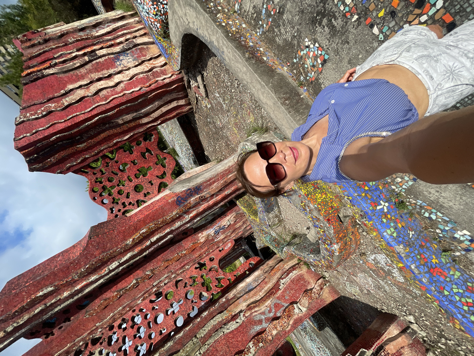 A woman wearing sunglasses and a blue striped shirt takes a selfie in front of a large, red, abstract mosaic sculpture. The sculpture is made of many red and white tiles and has holes cut out of it. The ground around the sculpture is also covered in colorful mosaics. The background shows a cloudy sky and some trees. The overall impression is an outdoor art installation.