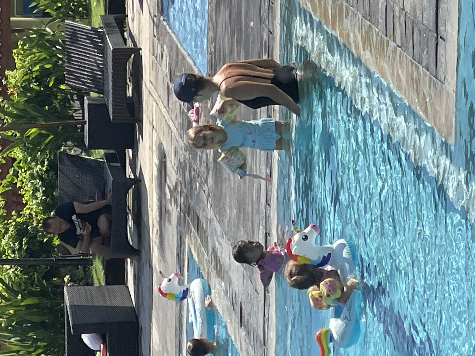 In Kuta Selatan, Indonesia, a resort pool area features a man in a black t-shirt and shorts seated on a dark gray lounge chair, looking at his phone. Several children are in the pool, some with inflatable unicorn pool floats. One young girl in a light blue swimsuit with yellow arm floaties is interacting with a woman wearing a black swimsuit and a navy blue baseball cap. Another child in a pink top is partially submerged, and a child with blonde hair is on a unicorn float. The pool is surrounded by a tiled deck and lush green foliage.
