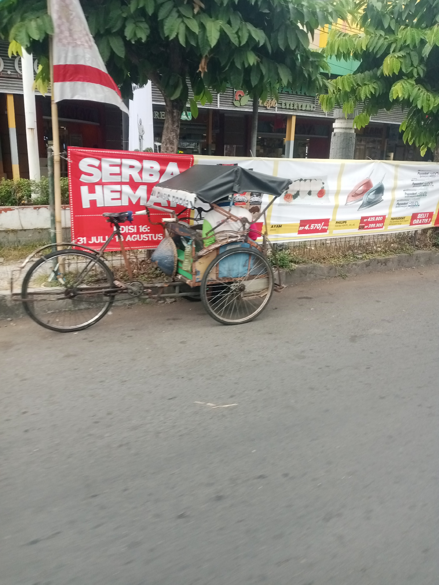 The image depicts a side view of a traditional Indonesian becak, a three-wheeled bicycle-powered vehicle, parked on the side of a paved road in Tasikmalaya, Indonesia. The becak has a covered passenger area with a black canopy and a blue seat. It is positioned in front of a large red banner that reads "SERBA HEMAT" (meaning "Very Economical") with the dates "31 JULI - 11 AGUSTUS" (July 31 - August 11) displayed below it, suggesting a promotional event. Behind the becak and banner, there are trees with lush green leaves and parts of buildings, indicating an urban or semi-urban setting. The road surface appears to be asphalt, with some wear and tear visible. The overall lighting suggests it is daytime, possibly late afternoon given the shadows. The image is taken from a slightly elevated position, looking downwards towards the becak.