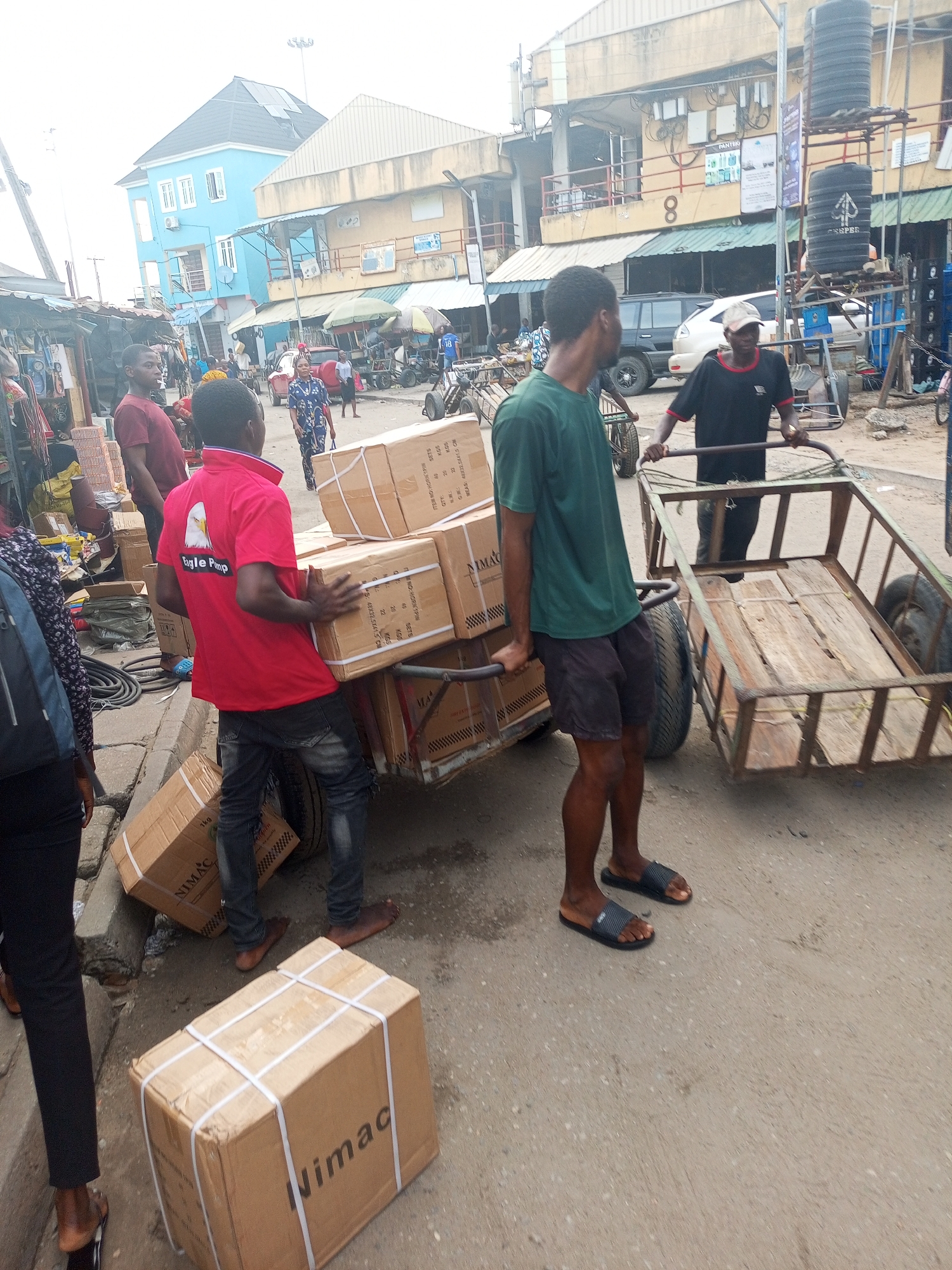 The image depicts a busy street scene in Satellite Town, Nigeria, bustling with commercial activity under bright daylight conditions. Several men are engaged in transporting goods using handcarts. In the foreground, a man in a red t-shirt, emblazoned with "Eagle Pimp" (likely "Eagle Pump") and an eagle logo, is carrying multiple cardboard boxes while another man in a green t-shirt pushes a loaded handcart. More "NIMAC" branded boxes, some with details like "ITEM NO: HOORNTPIN", "QTY", "SETS", "KGS", and "MEAS: 48X32.5X41.5", are visible on the cart and ground. To the right, another man in a white cap and black t-shirt pulls an empty, rustic handcart.
The setting is an urban commercial area, characterized by multi-story buildings, market stalls, parked vehicles (including cars and motorcycles), and various pedestrians. A distinctive blue building stands out on the left, while a tan building with the number "8" is visible further down the street. Overhead, power lines and a light pole are noticeable. Signs for "PANTEK" and "COSPER" (on water tanks) are visible in the background, along with a shirt bearing the number "18". The road surface is a mix of asphalt and dirt, and the overall impression is one of industrious activity and daily commerce. People appear focused on their tasks, reflecting a typical busy market environment.