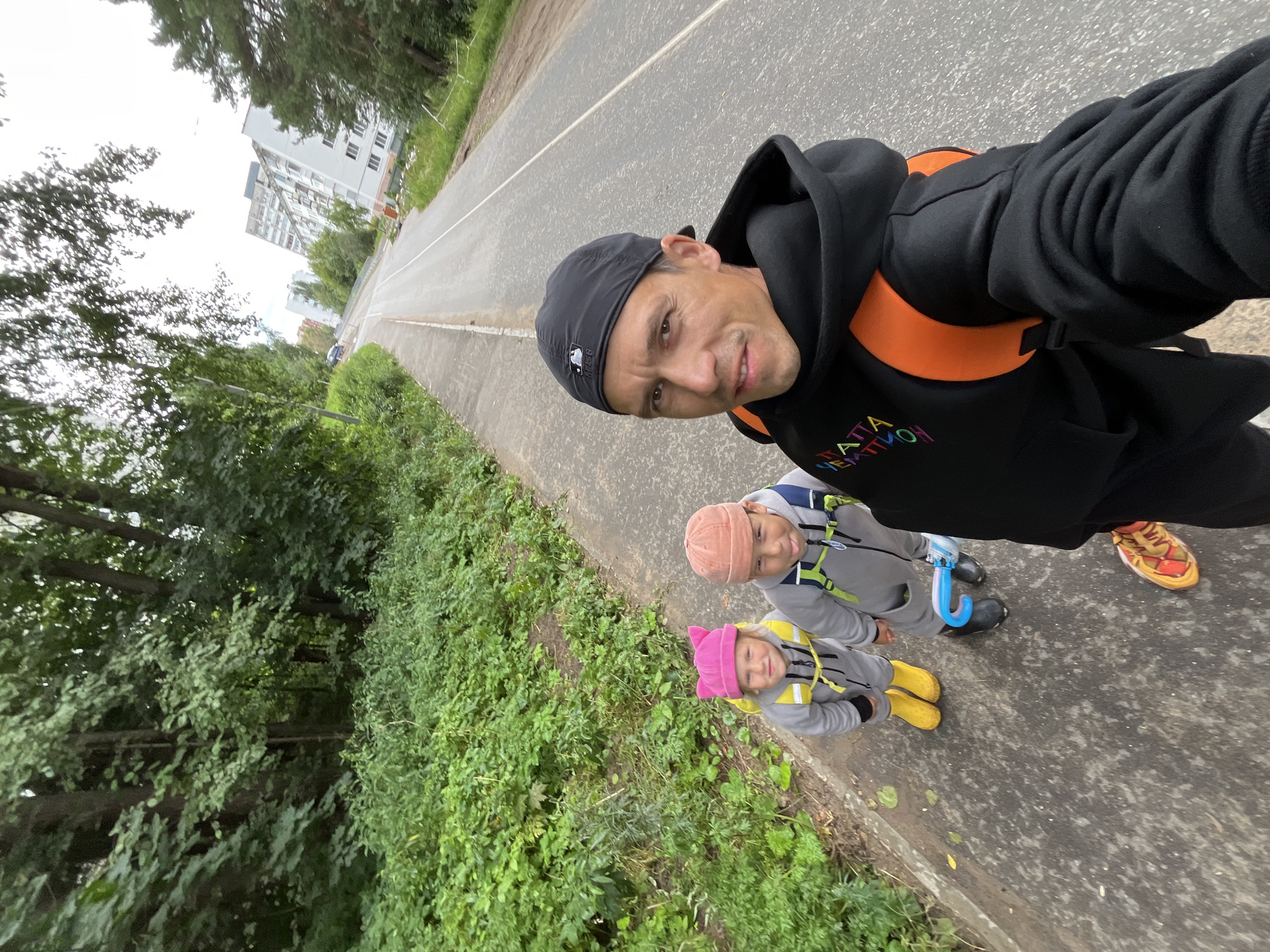 A man wearing a black baseball cap and black hoodie takes a selfie while walking on a paved path alongside a road. Two children, a girl in a pink hat and yellow boots and a boy in an orange hat and black boots, walk ahead of him on the same path. The girl wears a gray jumpsuit with a yellow backpack, and the boy wears a similar gray jumpsuit with a blue backpack. The path is bordered by lush green vegetation on the left and a road on the right. In the background, apartment buildings and trees are visible under a cloudy sky. The scene appears to be in the city of Chaikovsky, Russia, based on the architecture and general environment. The man is smiling at the camera, while the children are looking ahead. One of the children is holding a blue and white object, possibly an umbrella or toy.