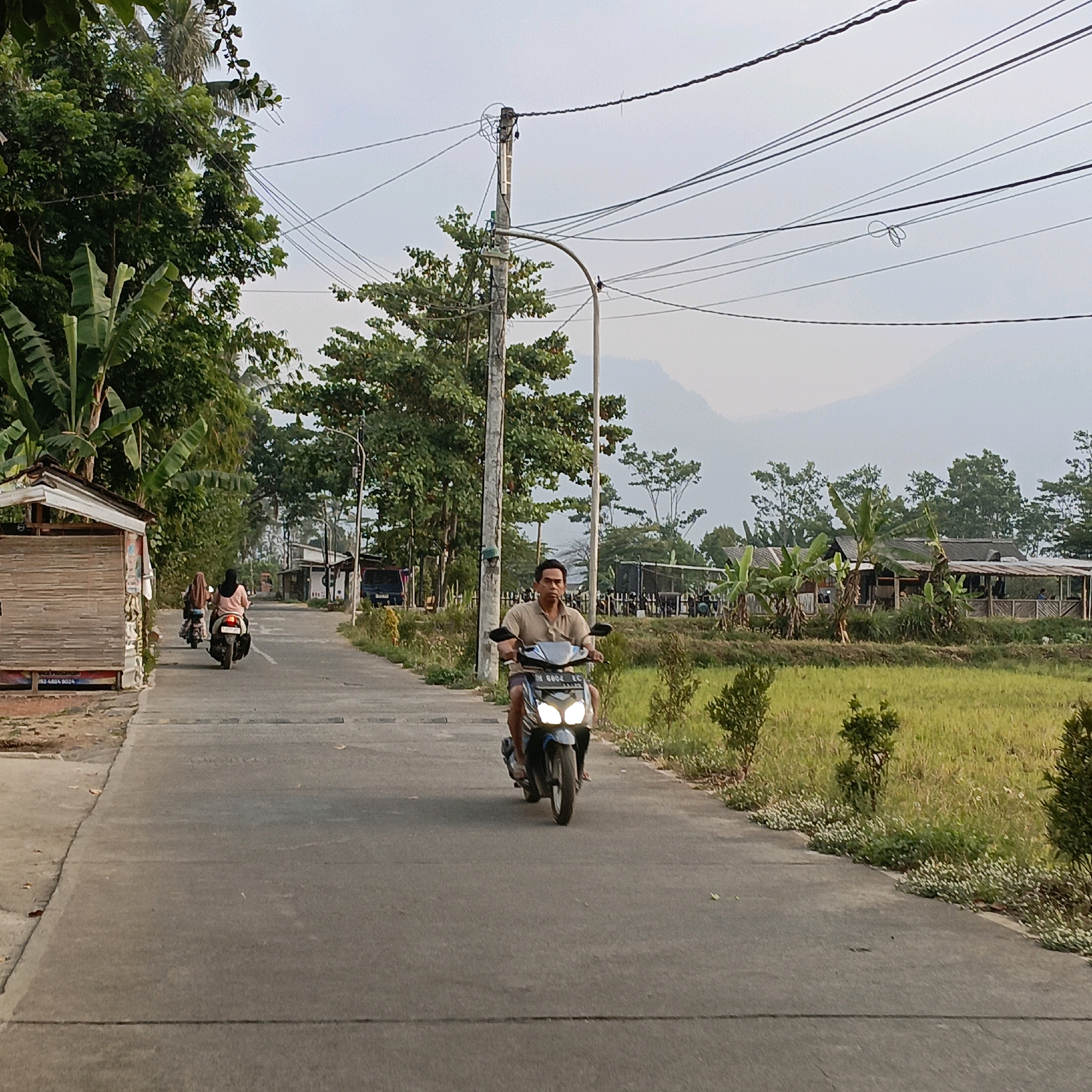 A motorcycle rider, a man wearing a light brown shirt and dark shorts, is riding a dark-colored scooter on a paved road. The scooter has its headlights on. The man is looking forward. In the background, another motorcycle with two riders is further down the road. To the left of the road, there is a small wooden structure with a thatched roof and bamboo siding, and beyond that, lush green trees. To the right of the road, there is a field of green grass and some small trees and bushes. In the distance, there are buildings with roofs and more trees. The sky is overcast, and in the far distance, mountains are faintly visible. The scene appears to be a rural or semi-rural area, possibly in Kesongo, Indonesia, given the presence of tropical vegetation and the style of the buildings and vehicles.