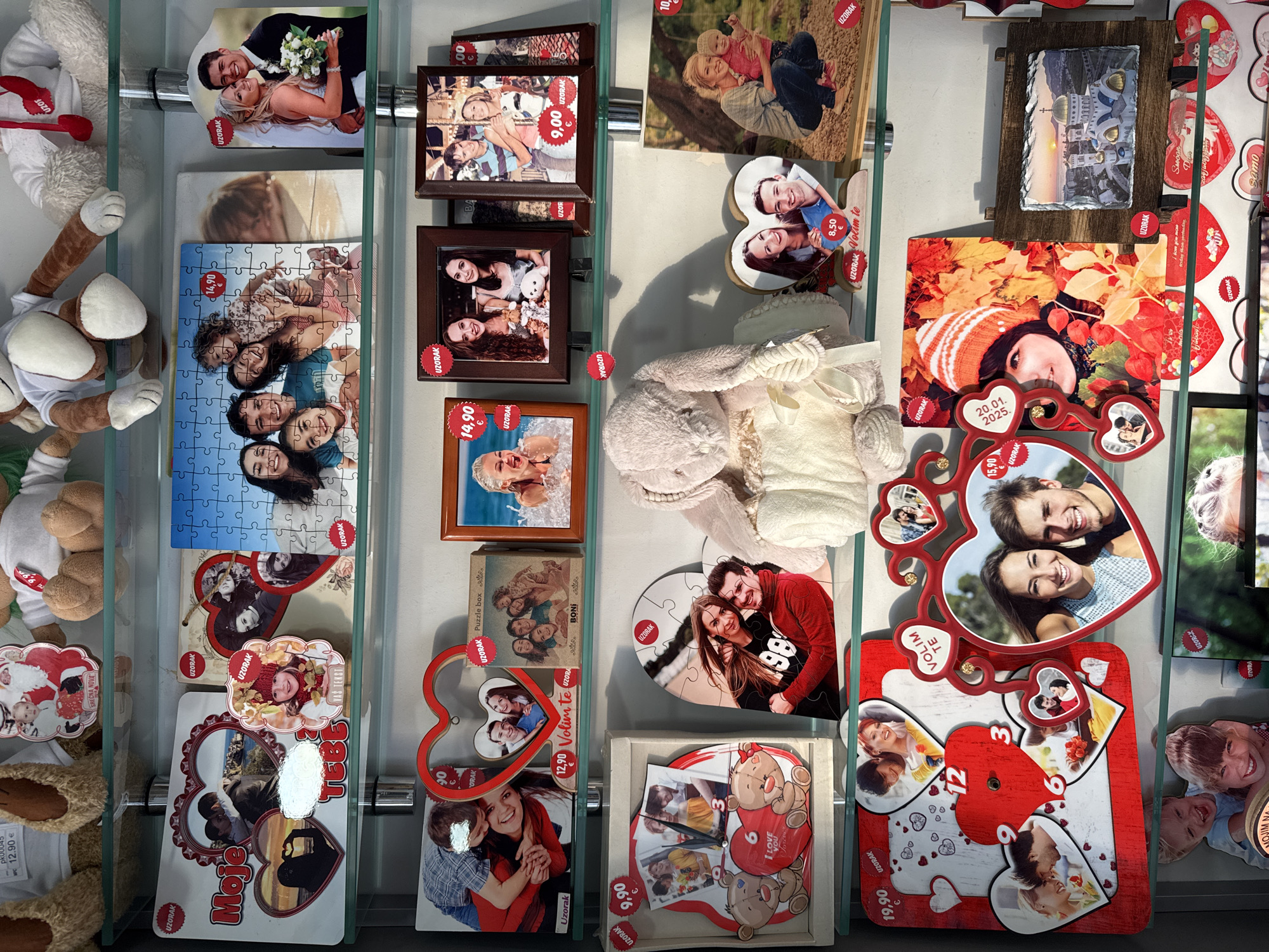 The image captures a display case, likely in a shop in Bar, Montenegro, featuring a variety of personalized photo products and plush toys. The products include photo puzzles, framed photos, photo clocks, and other novelty items with personalized images. Several of the products bear the word "UZORAK" and price tags in Euros. The toys are mainly plush animals. The photo products feature pictures of individuals and couples. The environment appears to be well-lit.