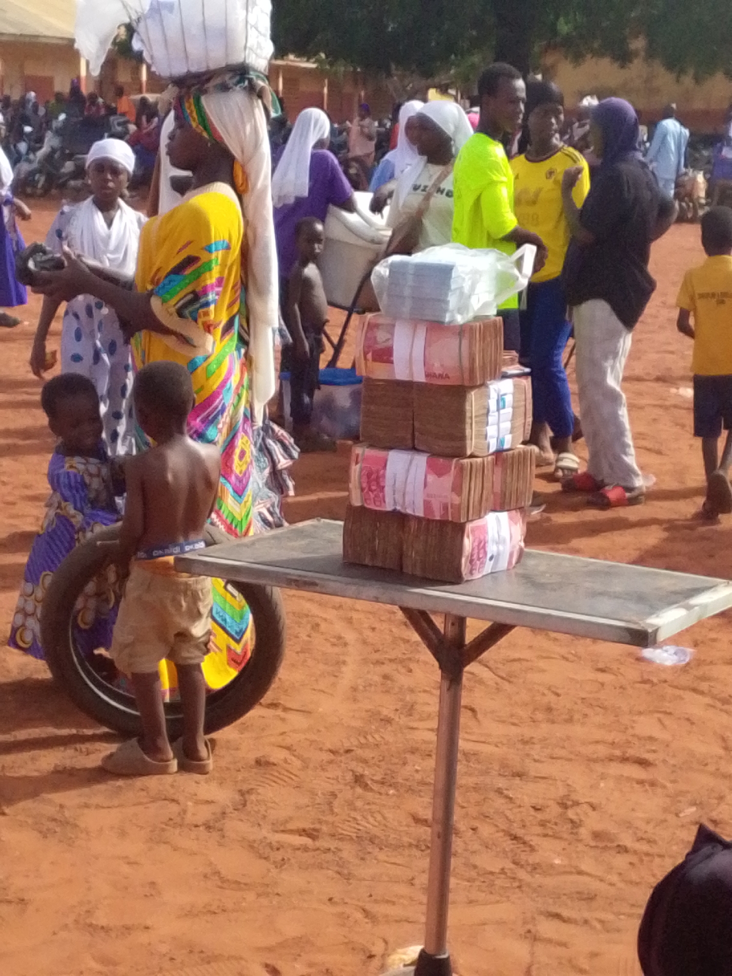 The image shows an outdoor scene, likely in Tamale, Ghana, with a sandy ground surface. At the center is a metal folding table supporting four stacks of what appear to be bound bundles of Ghanaian cedi notes, some labeled "GHANA". Two children are beside the table; one shirtless and wearing shorts stands next to a tire leaned against the table's leg, while another, dressed in a patterned blue dress, looks on. A woman in a yellow and colorful wrap carries a load on her head to the left of the table. In the background, various adults and children are seen, some wearing Muslim headscarves, and some in colorful clothing. Some people are standing in small groups engaging with each other. One group in the middle of the background appears to be interacting with each other.