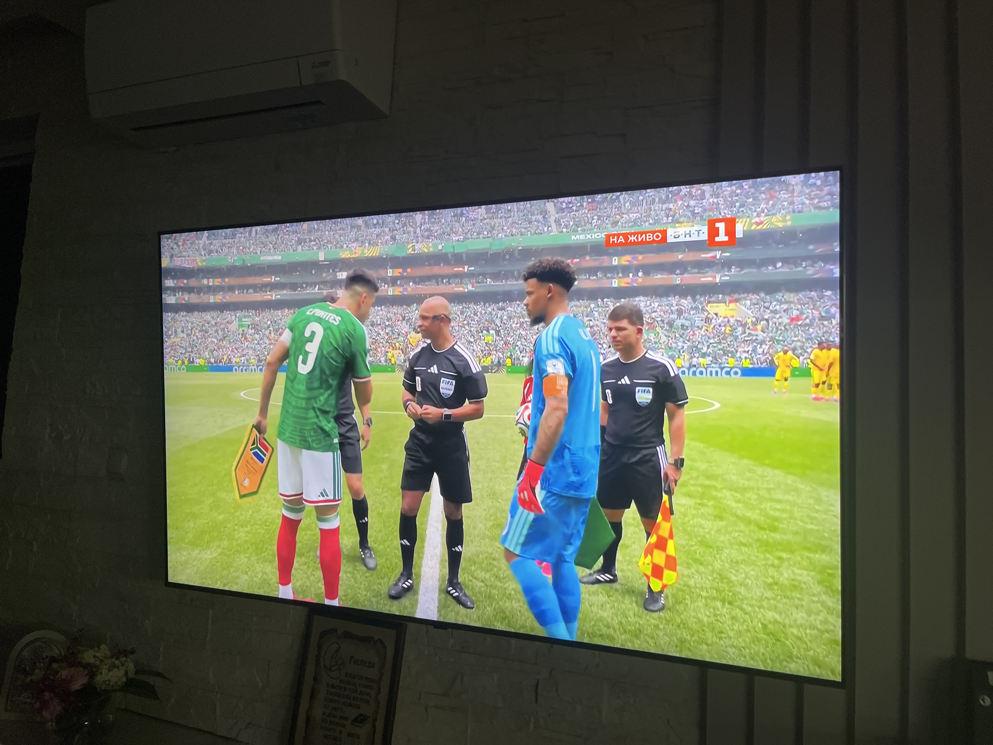 The image displays a television screen showing a live broadcast of a soccer match. In the foreground of the broadcast, four individuals are visible on a green soccer field. Two are wearing green and white soccer uniforms with the number '3' and 'C. Fuentes' on the back of one. The other two are dressed as referees in black uniforms with "FIFA" logos. One referee holds a checkered yellow and red flag. In the background, a stadium filled with spectators is visible. A score overlay on the screen shows "MEXICO" and "НА ЖИВО 1-1". The television is mounted on a wall above a brick-like textured surface. To the left of the television, on a table, is a vase with flowers. To the lower right of the television, a framed piece of artwork or document is visible. The scene appears to be indoors, in Varna, Bulgaria.