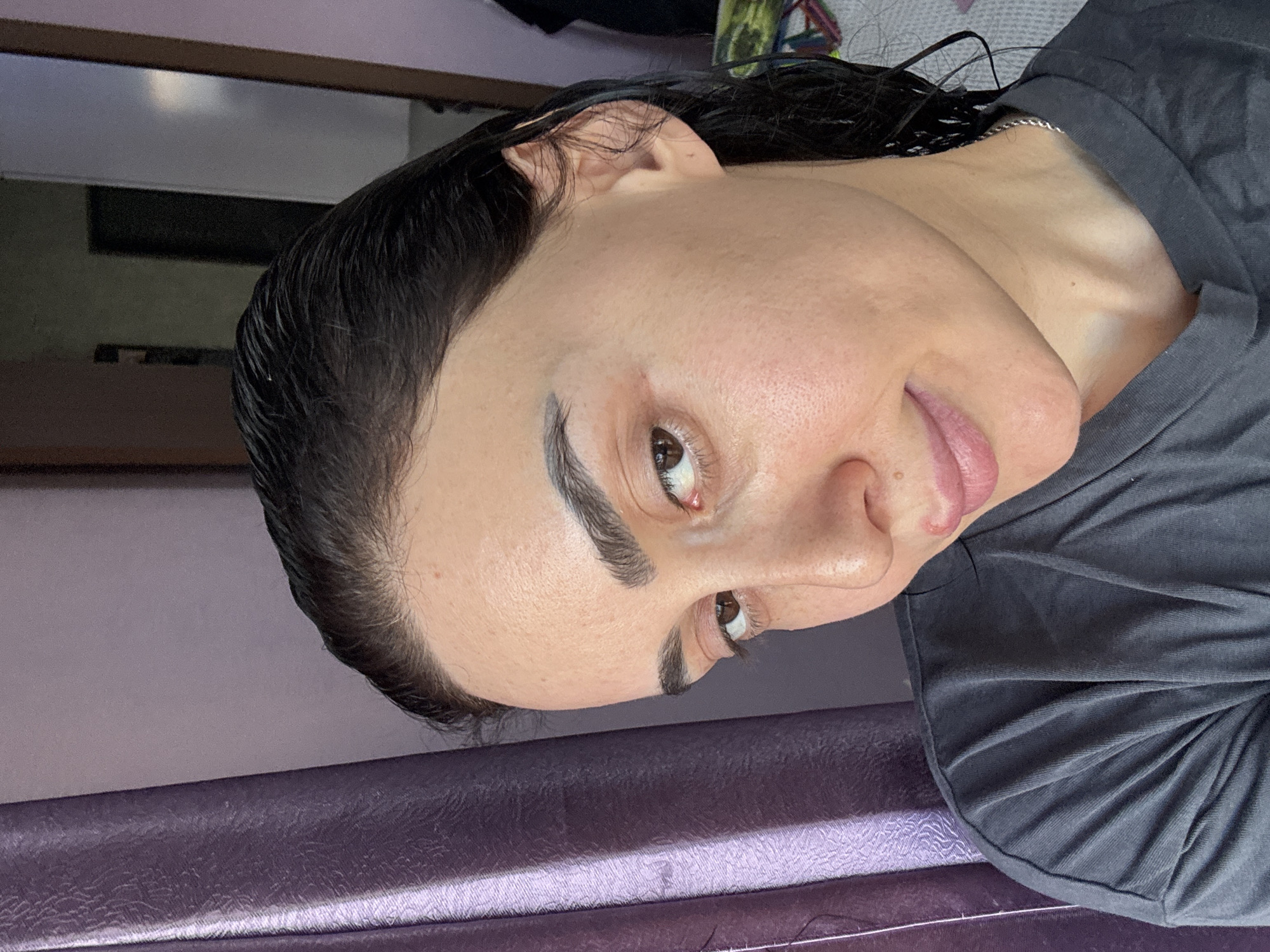 A close-up, sideways portrait captures a person, likely female, with dark, slicked-back hair that appears wet. Her face is turned towards the viewer, featuring well-defined, dark eyebrows and brown eyes. She has fair skin with visible pores. Her expression is a subtle smile or smirk, with the tip of her tongue slightly visible, conveying a relaxed or playful mood.
The person is wearing a dark grey t-shirt or top, and a thin silver chain is visible around her neck. Part of her ear is visible, tucked behind her hair.
The setting is indoors. A prominent purple upholstered surface, possibly a seat or bench with a textured pattern, occupies the foreground and bottom portion of the image. The background is out of focus, revealing blurry elements that suggest a room interior, including what appears to be a window reflecting light and darker, indistinct shapes. The lighting suggests indoor illumination, possibly complemented by natural light, but the time of day is not clear. There is no visible text. While the location is contextualized as Tbilisi, Georgia, the image itself does not contain specific visual cues to confirm this.