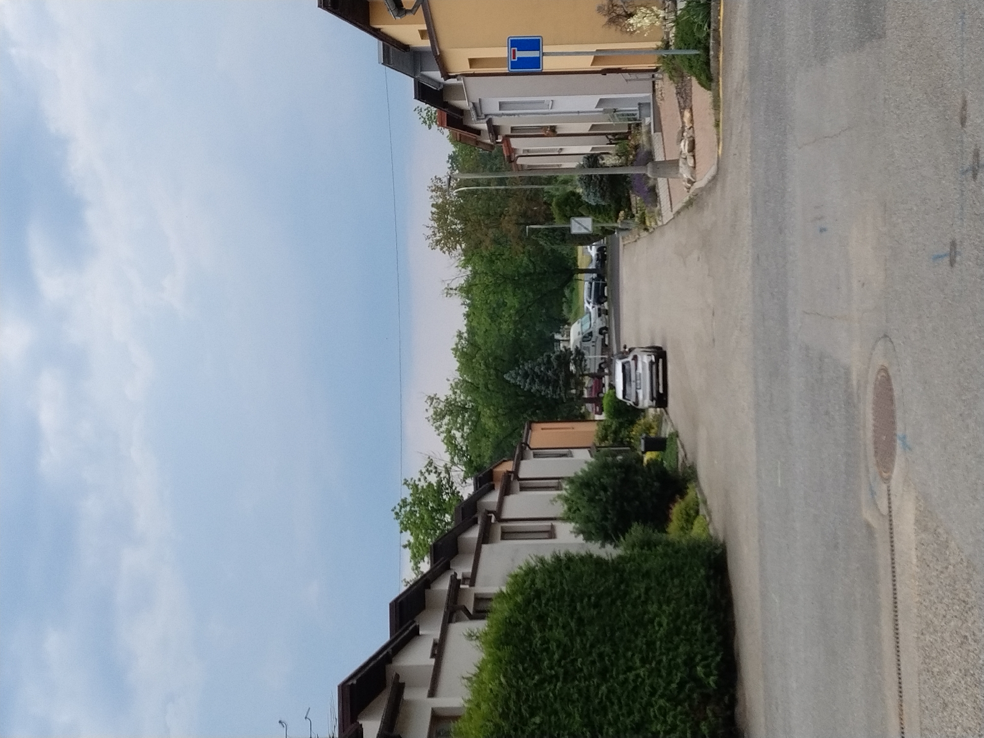 The image depicts a residential street in Hluboká nad Vltavou, Czechia. On the left side of the street, several attached white houses with dark brown roofs and gutters are visible, bordered by dense green hedges. A white car is parked on the street in front of these houses. The right side of the street features more attached houses, one with a yellow facade and a dark roof, another white house, and further down, houses with a light beige facade. A blue and white traffic sign indicating a dead end is posted next to the yellow house. The street is paved with asphalt, and a drainage grate runs along the edge of the sidewalk. In the distance, trees line the street, and several vehicles, including a white camper van and a dark car, are parked further down the road. The sky is cloudy with patches of blue.