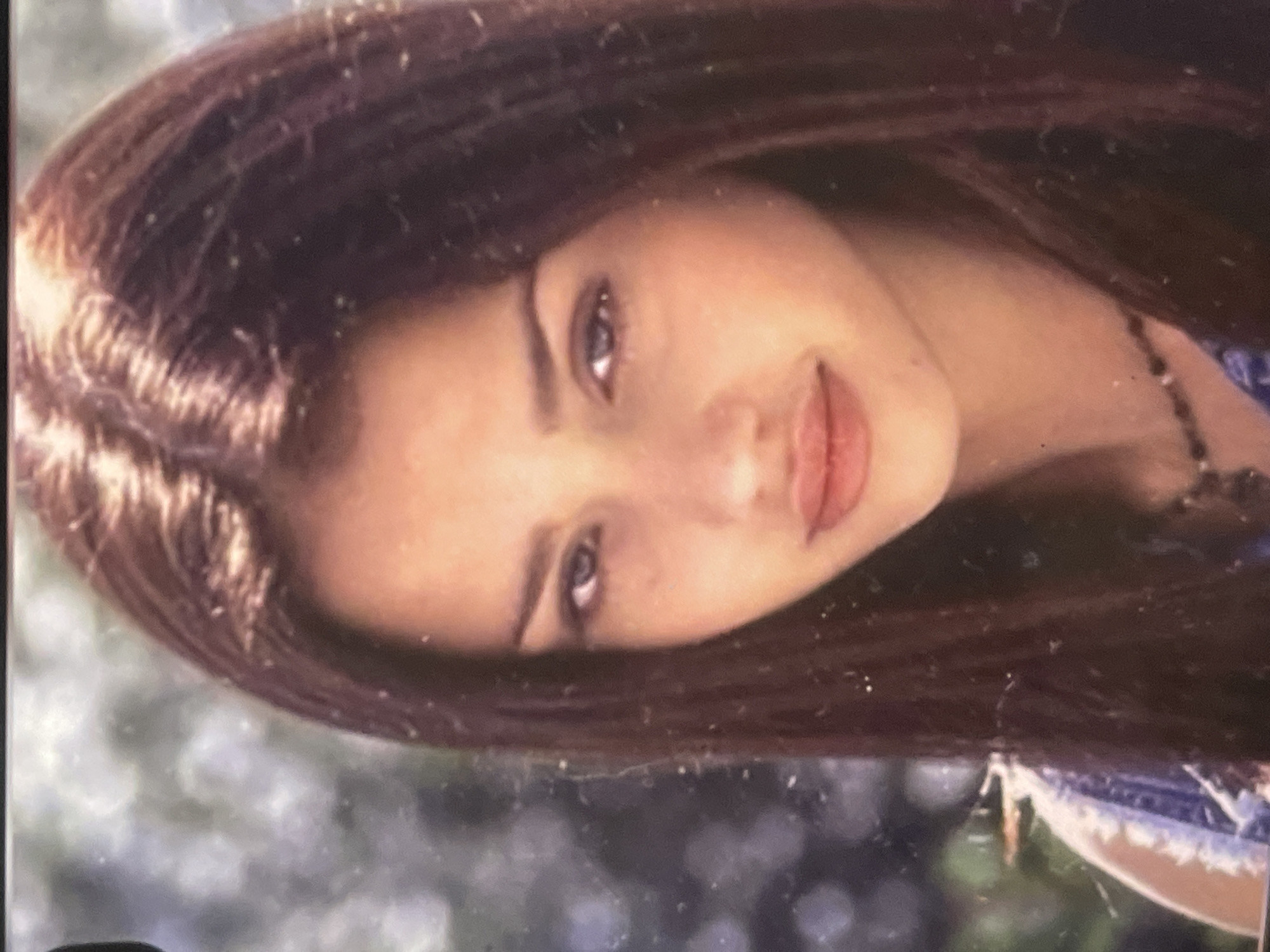 A portrait of a young woman is presented, rotated 90 degrees clockwise. She has long, dark brown hair that cascades around her face. Her head is tilted slightly, and she looks directly at the viewer with a calm expression and a subtle, gentle smile. Her lips are a natural pink hue, and her eyebrows are dark and well-defined. A dark, possibly beaded, necklace is visible on the right side of her neck. The background is softly blurred with bokeh effects, showing hints of green and lighter tones, indicative of an outdoor setting such as a park or garden during daylight hours. The lighting appears natural and soft. The image exhibits some graininess and minor dust specks, suggesting it may be a scanned photograph. No text is visible. The scene portrays a serene moment in an outdoor location, potentially in Poltava, Ukraine.