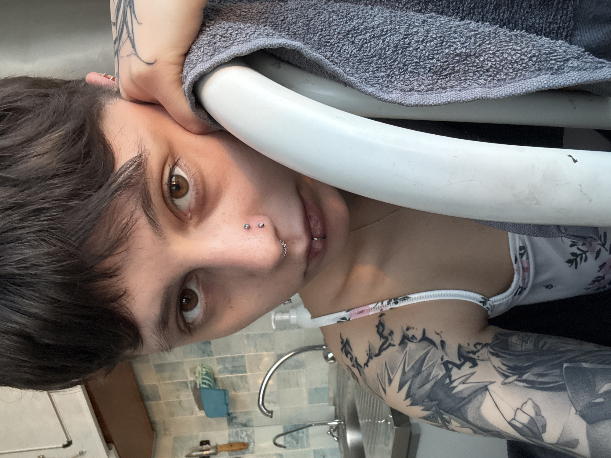 A close-up, slightly tilted portrait shows a person with short, dark hair and brown eyes looking directly at the camera. Their left arm is bent, and their hand holds a grey terrycloth towel draped over a white, curved object, possibly a chair back or a drying rack. The person has several facial piercings, including two studs on their right nostril, a small hoop on their left nostril, and a stud piercing on their philtrum (above the upper lip). An earring is also visible in their left ear.
They are wearing a white top with a delicate floral pattern and a ruffled elastic trim. A detailed sleeve tattoo is prominent on their right arm, depicting several figures in what appears to be an anime or manga art style. The background suggests an indoor setting, likely a bathroom or kitchen, with light blue and grey patterned tiles on the wall. A chrome faucet and sink are visible, along with a blue sponge and a wooden-handled utensil. No visible text is present. The context provided is Saint Petersburg, Russia.