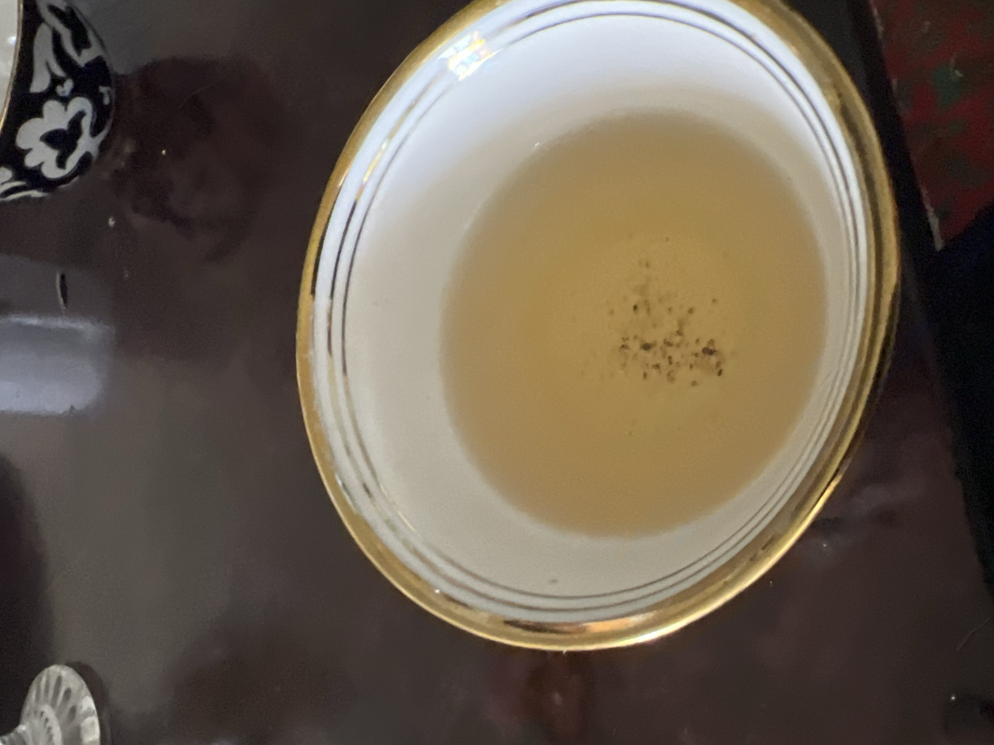 A close-up, high-angle shot shows a white bowl with a gold rim filled with a light-colored liquid, possibly tea. The liquid has a few small dark specks floating on its surface. The bowl is placed on a dark, reflective surface, likely a wooden table, which occupies most of the frame. In the upper right corner, out of focus, is another dark bowl with a white floral pattern. The overall lighting is dim, and the focus is on the bowl of liquid. The image is a still life, with no people or action present. The location is Hazorasp, Uzbekistan.