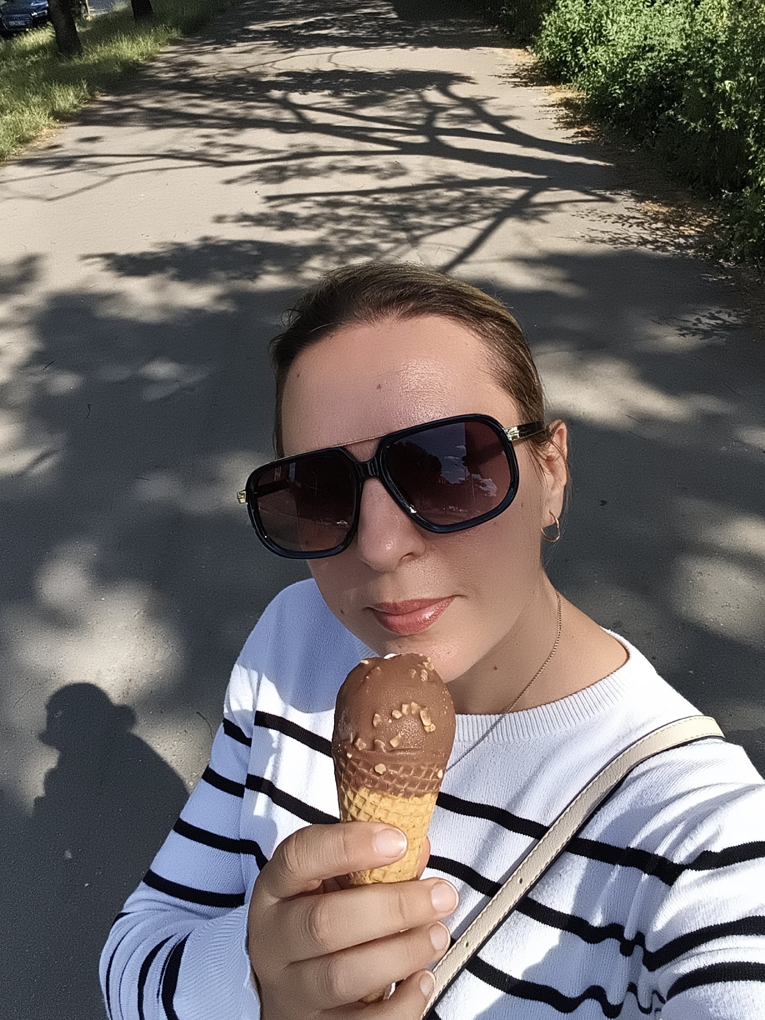 In Wrocław, Poland, a woman is captured in a selfie, holding a chocolate ice cream cone in her right hand. She wears large, dark-lensed sunglasses and a white sweater with black stripes. A thin gold chain is visible around her neck. The background is an asphalt path shaded by trees, suggesting an outdoor setting possibly in a park or along a tree-lined street. The sun casts strong shadows of tree branches onto the path.