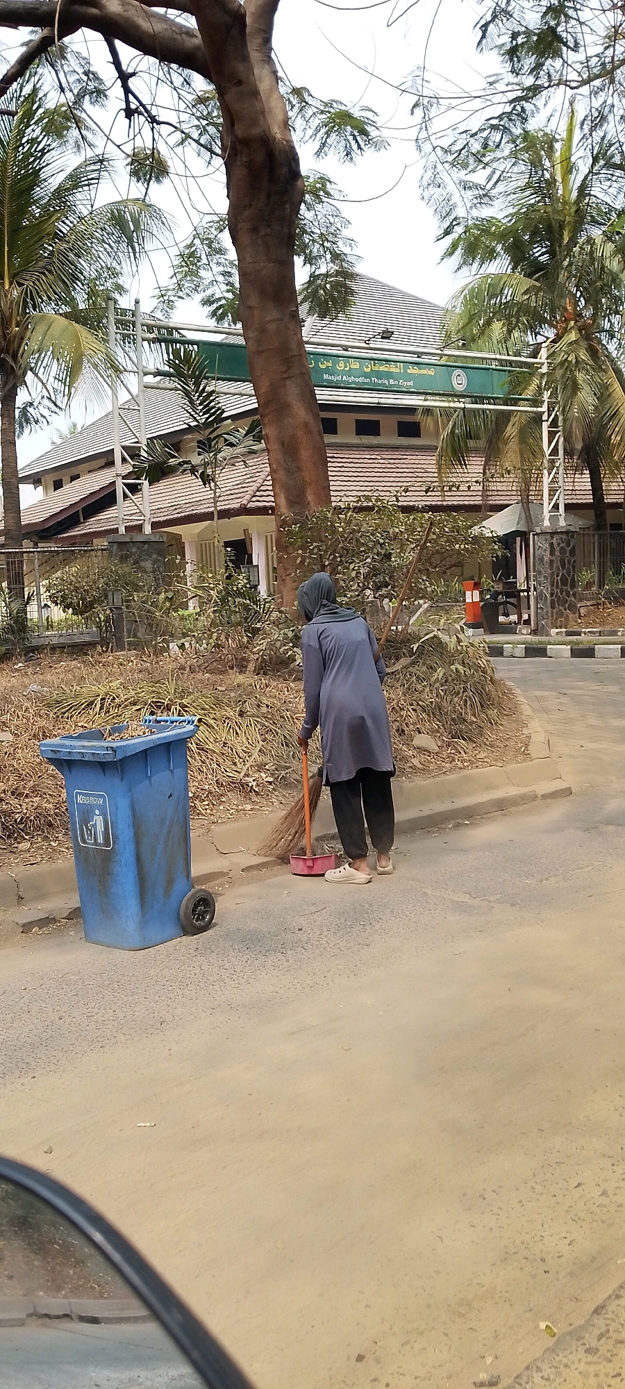 A person dressed in a gray tunic and black pants, wearing white slip-on shoes, is sweeping the dusty road with a broom. The person's head is covered with a gray hijab. To the left of the person is a large blue trash can with the label "KALEBOW" and a recycling symbol. The trash can has a broken lid and appears to be full of debris. In the background, there is a building with a tiled roof, flanked by palm trees and other foliage. A green sign above the building reads "Masjid Alghodfan Thariq Bin Ziyad" in Arabic and English. The scene appears to be in Karangsatria, Indonesia, given the context.