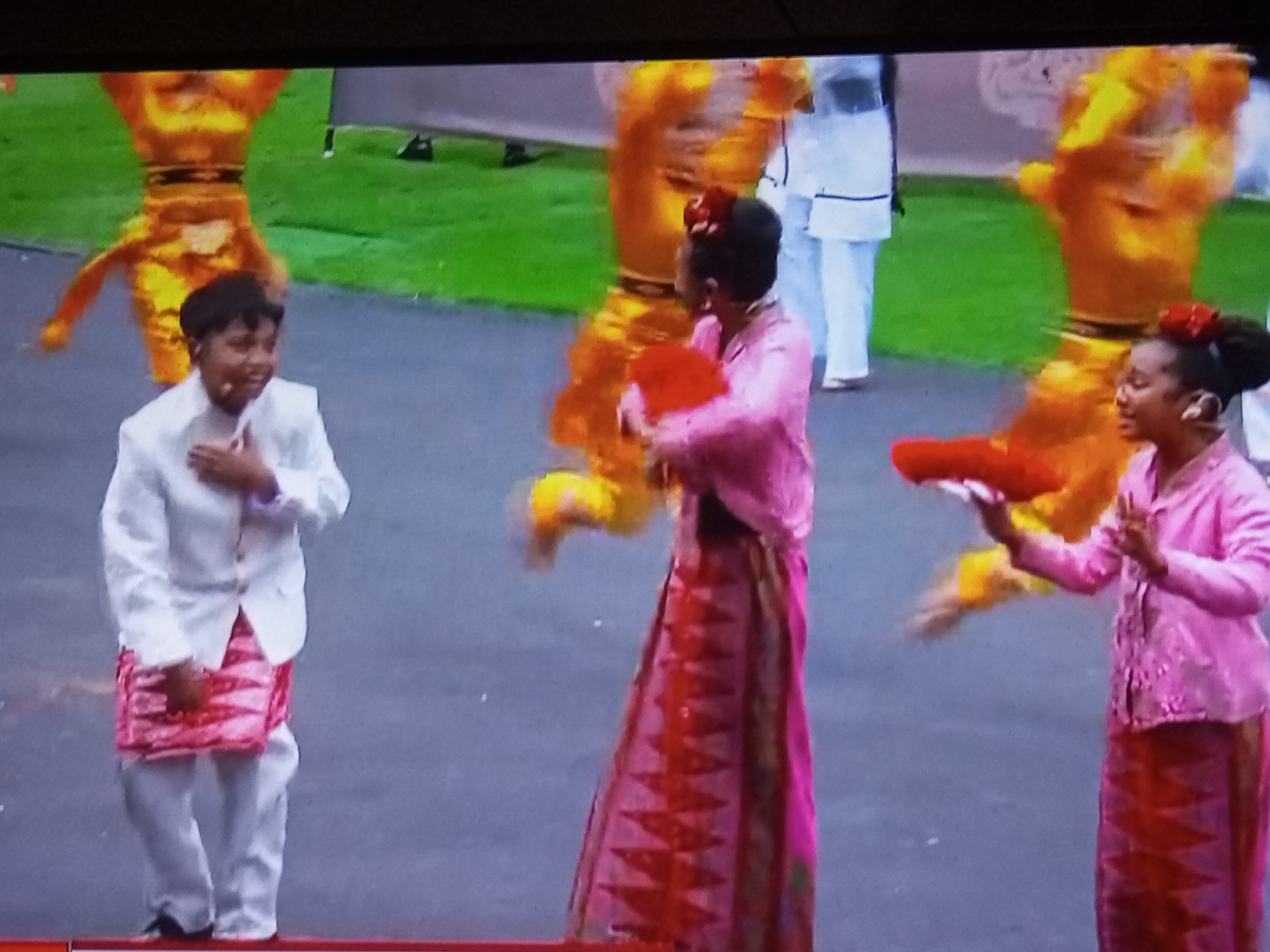 The image depicts a scene with several individuals dressed in traditional attire, likely during a cultural performance or festival. In the foreground, a young boy in a white jacket and patterned pink and white sarong stands with his hand on his chest, looking to his right. To his right, a person in a vibrant pink outfit with a patterned red and pink skirt is holding a red object. Further to the right, another individual, also in a pink outfit with a patterned skirt, is holding a red object. In the background, two figures in bright orange, elaborate costumes are blurred, suggesting movement. The ground appears to be paved, with a grassy area behind the performers. The overall impression is of a lively, celebratory event. The location is identified as Banyudono, Indonesia.