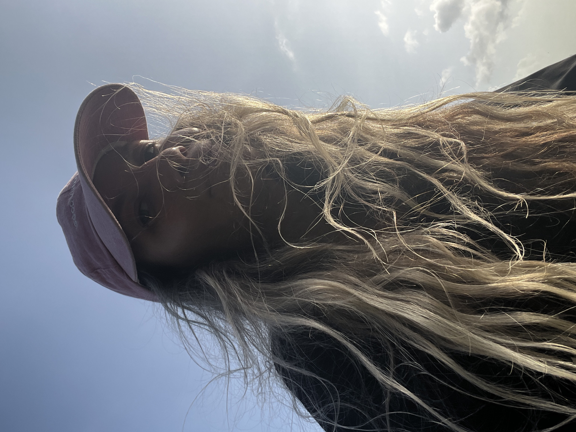 A low-angle, close-up selfie shows a person wearing a pink baseball cap and a dark top against a clear blue sky with scattered clouds. The person's long, blonde hair is windswept across their face, partially obscuring their features. The sun is shining from the right, creating a bright halo effect around the hair and highlighting the texture of the strands. The image appears to have been taken outdoors, with the sky dominating the background. The context of Dondra, Sri Lanka is not discernible from the visual information provided in the image itself.