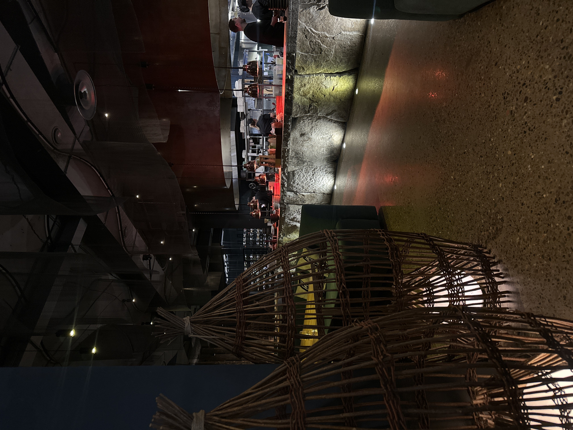 The media file captures the interior of what appears to be a restaurant or bar in Krasnoyarsk, Russia, characterized by its dim, atmospheric lighting and distinct design. The image is oriented vertically.
In the foreground, dominating the lower-right section, are two large, elongated decorative elements resembling woven baskets or traps, crafted from dark brown reeds or slender wood. These structures house internal light sources that emit a soft, diffused glow.
The midground features a prominent bar or counter area running horizontally. The wall beneath this counter has a rough, textured, stone-like appearance, illuminated by several embedded spotlights casting warm yellow and red hues. To the right of this wall lies a dark, speckled floor, reflecting the ambient light.
Behind the counter, in the mid-left portion of the image, several people are visible. One individual stands tall, likely a bartender, while others are seen further down the counter, engaged in work or conversation. More indistinct figures are scattered in the background.
The upper-left section of the image reveals a complex ceiling or upper wall structure, featuring exposed grilles, ducts, and multiple recessed spotlights that cast bright, focused light. Dark architectural panels, possibly metallic or wooden, complete this area. Dark upholstered seating is partially visible in the lower-left corner and near the bar.
The scene depicts an evening or nighttime setting, evident from the heavy reliance on artificial lighting to create a moody and sophisticated ambiance. The combination of natural textures like stone and woven reeds with modern industrial elements contributes to a unique aesthetic. No discernible text is visible within the image.