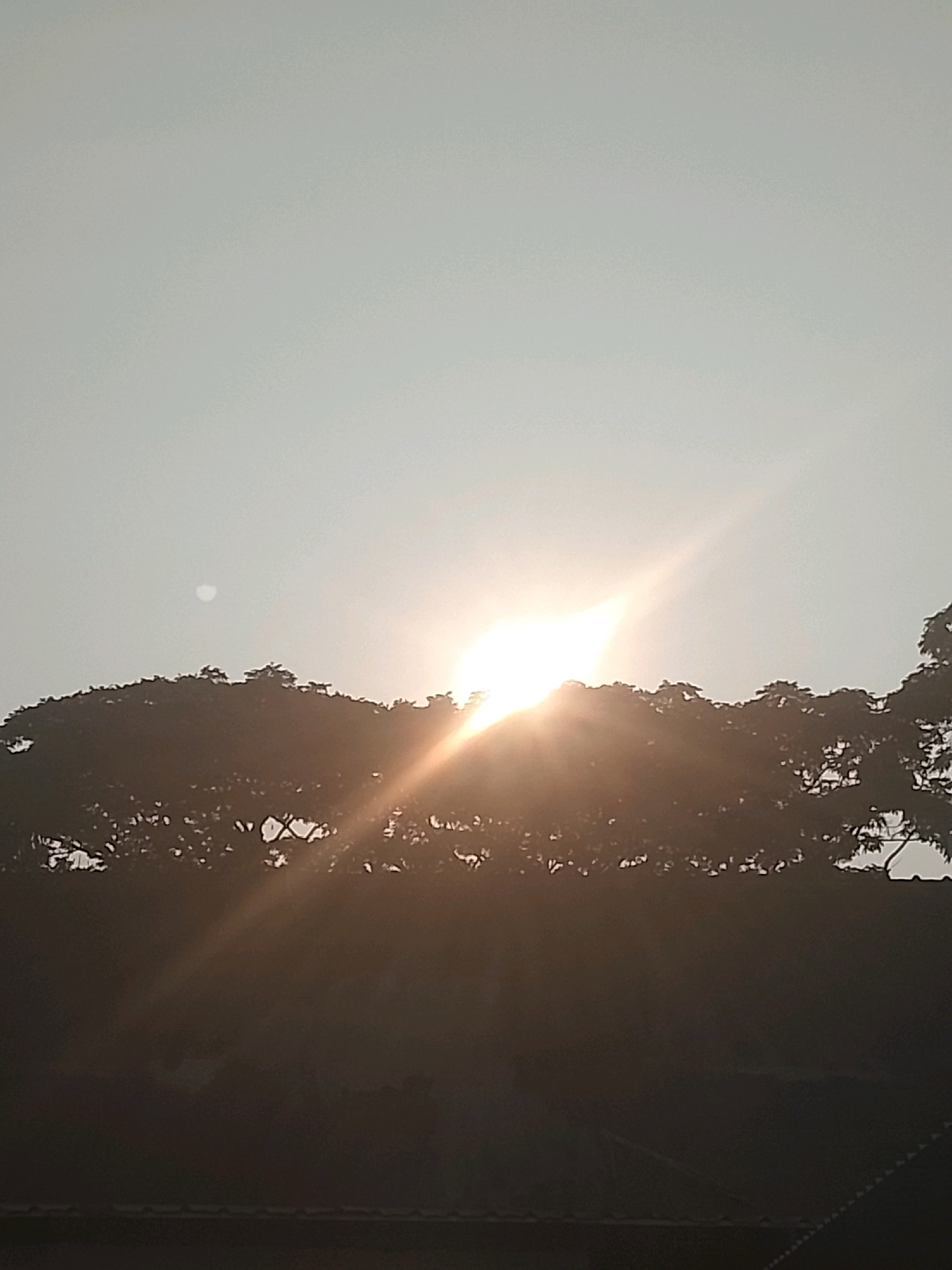 The image depicts a silhouetted line of trees against a bright sky. The sun is a strong, glowing orb positioned behind the trees, casting a lens flare that extends diagonally across the upper right portion of the frame. A faint, circular shape, possibly the moon or a very hazy sun, is visible in the upper left quadrant of the sky. The foreground is dark and indistinct, suggesting it is either late evening or early morning. The overall scene appears to be a natural landscape with the sun prominently featured. The location is identified as Slawi, Indonesia.