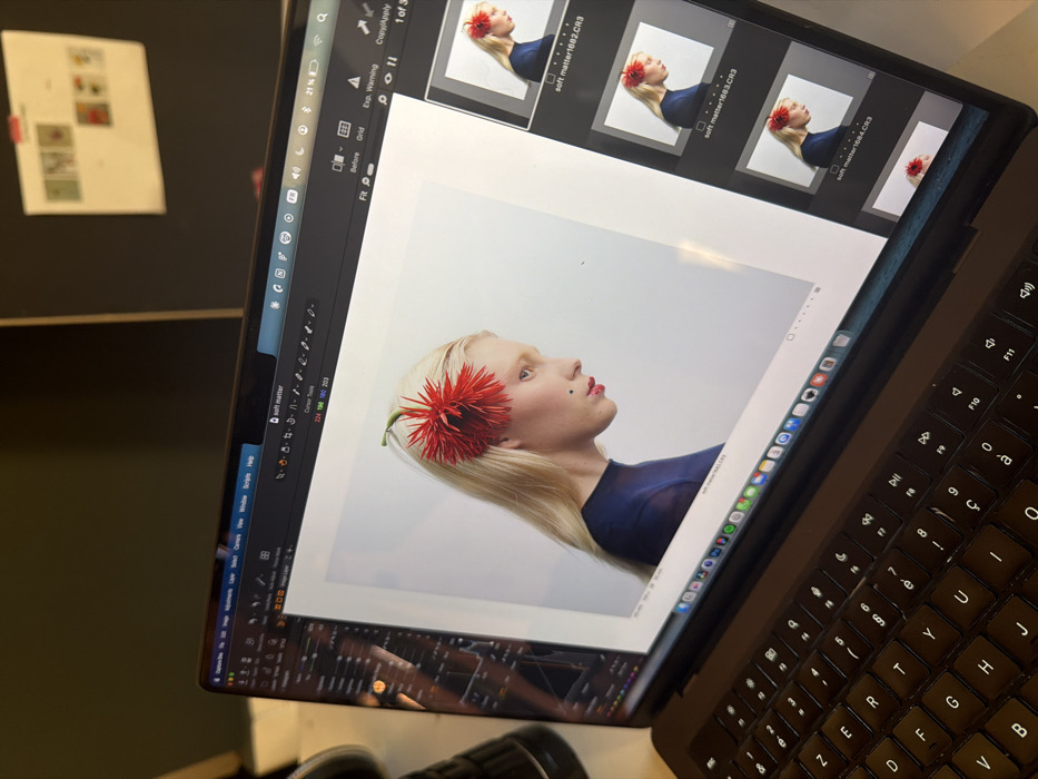 A dark-colored laptop, identified as a MacBook Pro with an AZERTY keyboard layout, is prominently displayed in the foreground, viewed from an oblique, elevated angle. The screen shows the Capture One professional photo editing software. The main display area features a profile photograph of an individual with long, straight blonde hair, light skin, and red lipstick, looking towards the right. A prominent, spiky red flower is affixed to the hair above the left temple. A small dark mole is visible on the cheek below the left eye. The individual is wearing a dark blue, semi-sheer, high-necked garment, and the background of the photograph is a plain white. A vertical filmstrip of thumbnail images, including files like "soft matter1682.CR3," occupies the right side of the screen. The macOS menu bar at the top indicates a 21% battery charge and other system icons. To the left of the laptop, a dark, cylindrical object, likely a camera lens, is partially visible. The background is dark and out of focus, with a light-colored sheet of paper bearing small images or notes, secured by a red clip, visible on a dark wall or board in the upper right. The AZERTY keyboard layout specifically indicates the scene's location is in Paris, France.
