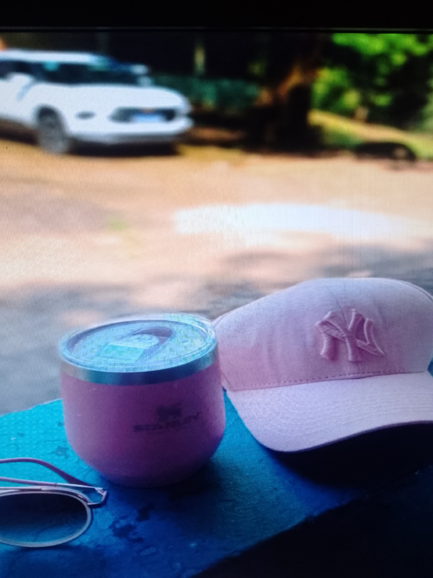 The image depicts a pink Stanley cup, a pink New York Yankees baseball cap, and a pair of sunglasses on a dark blue surface. In the blurred background, there is a white vehicle and some greenery, potentially trees. The location is unknown.