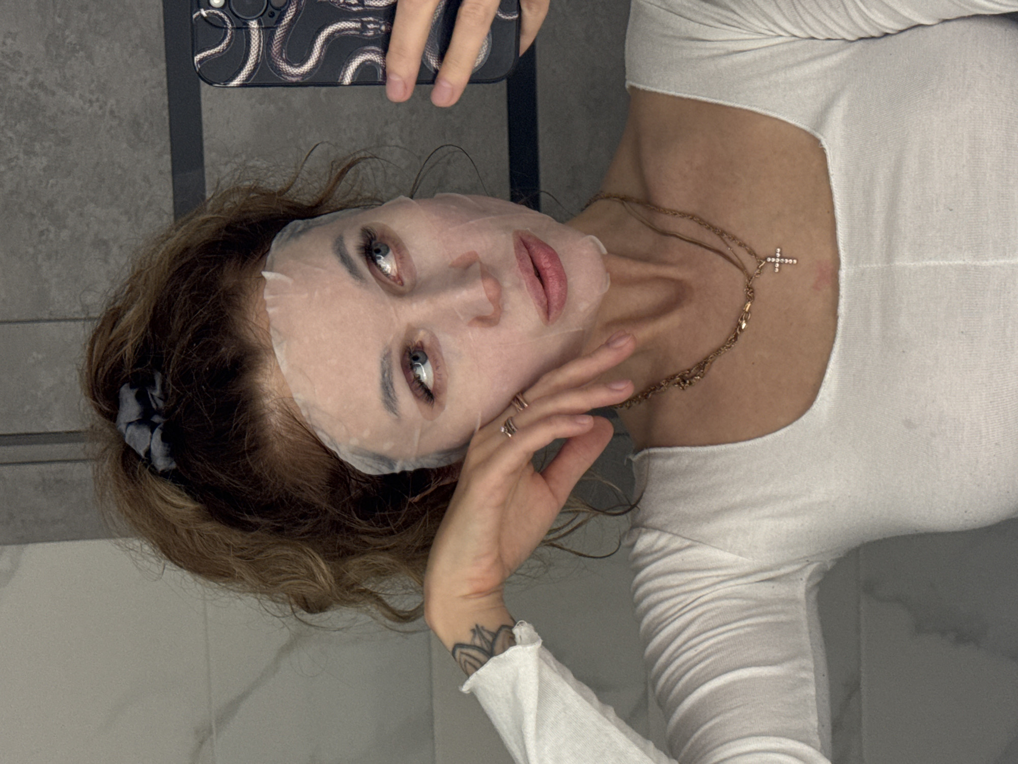 A woman with curly brown hair pulled up in a bun, wearing a white long-sleeved shirt and a sheet mask, takes a selfie in a bathroom. She is wearing two gold necklaces, one with a cross pendant, and several rings on her fingers. A tattoo is visible on her left forearm. The background appears to be a tiled wall. The city of Chaikovsky, Russia is not depicted in the image.