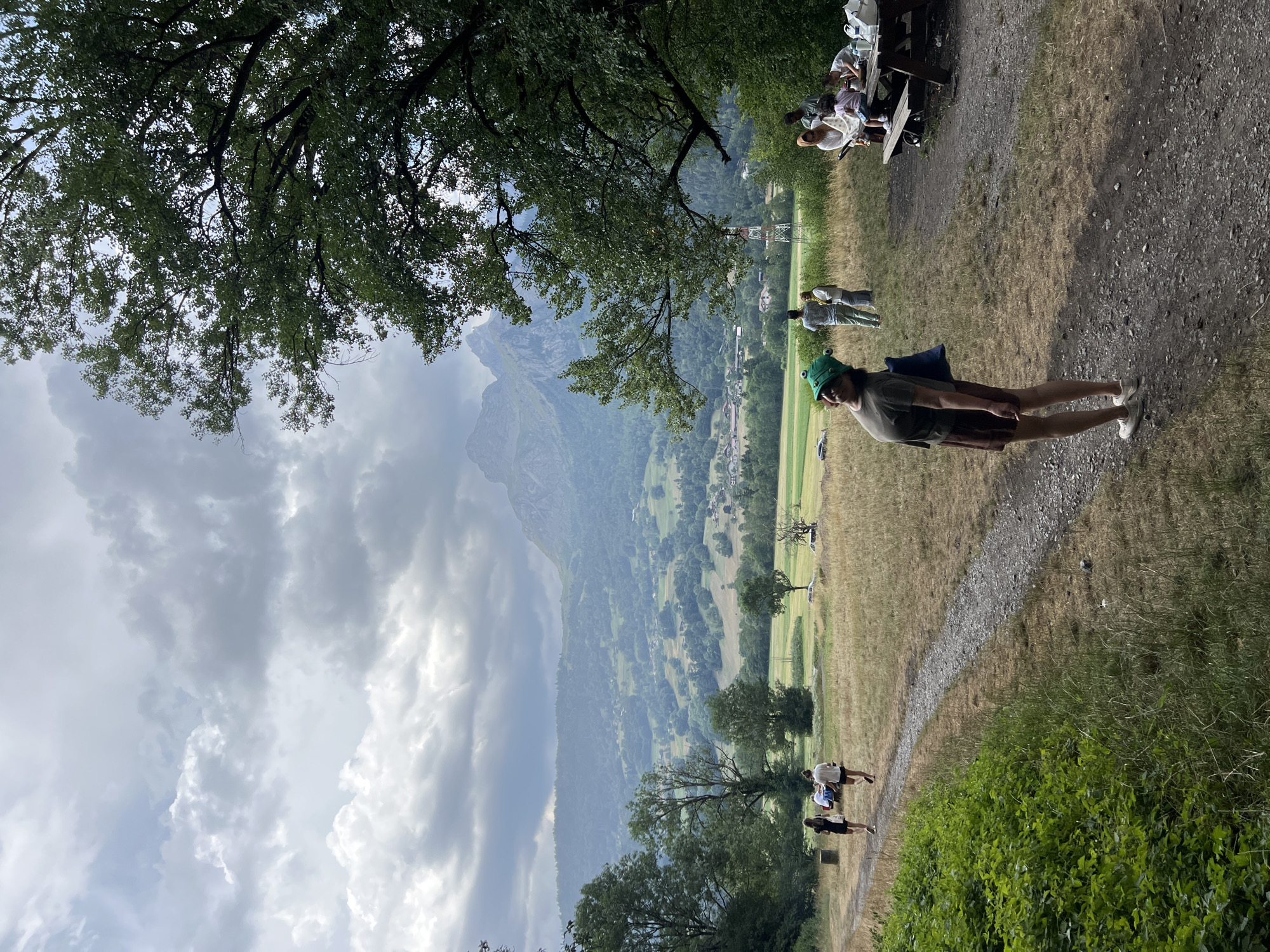 The image depicts a rural landscape with a young woman in the foreground wearing a green frog-shaped hat and a brown skirt. She is looking up and to her right, with her left hand at her side. Behind her, a gravel path winds through a grassy field towards a distant mountain range. Several people are visible in the mid-ground, some walking and others gathered around a picnic table. A village and trees dot the valley between the path and the mountains. The sky is overcast with dramatic clouds. The scene appears to be located in the outskirts of Sallanches, France, given the mountainous terrain.