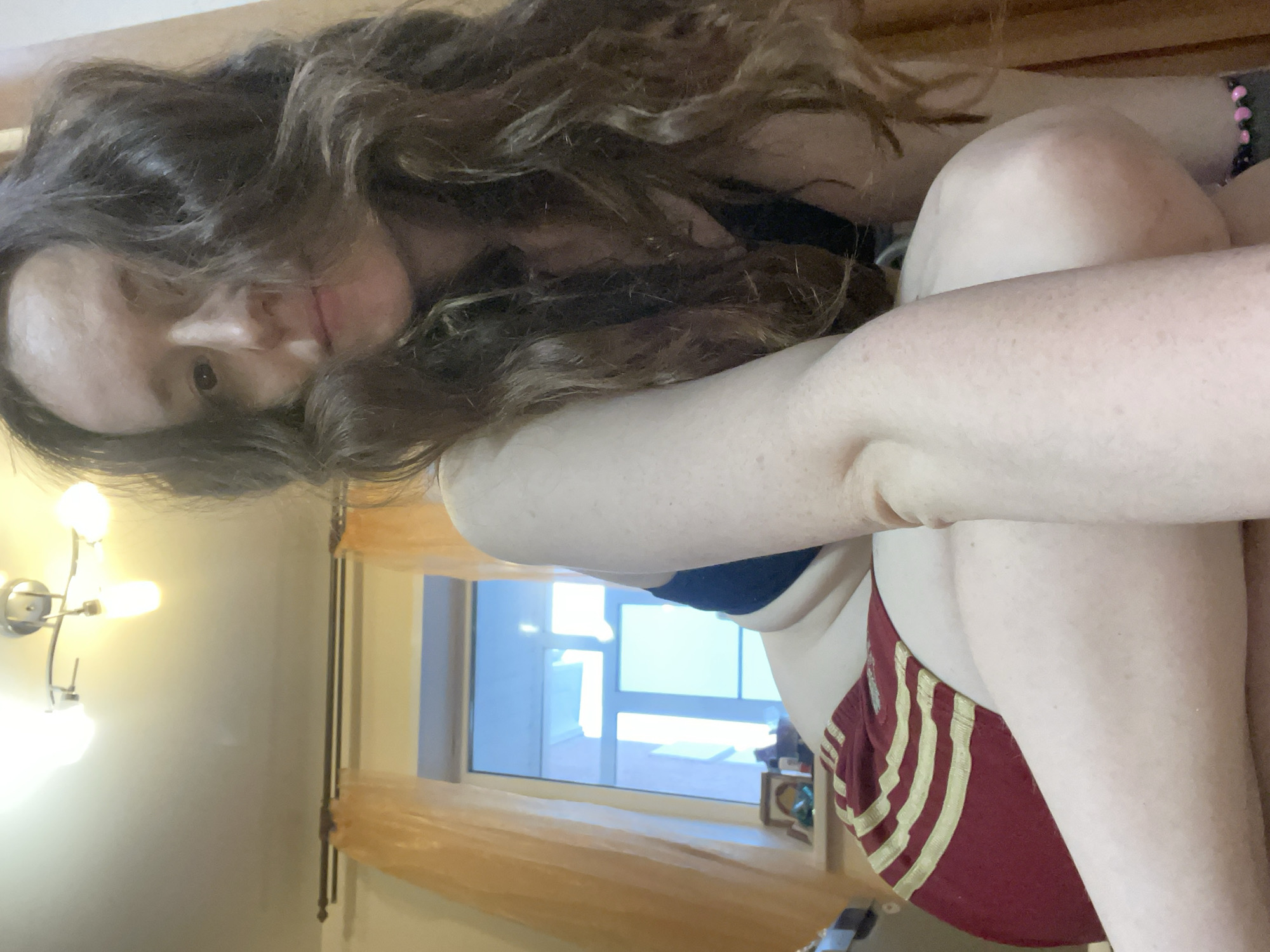 A person is in a room with a window and a sconce on the wall. The person has long brown curly hair. The person is wearing a blue sports bra and maroon shorts with gold stripes. The location cannot be identified as Usol'e, Russia, from the visual information provided.