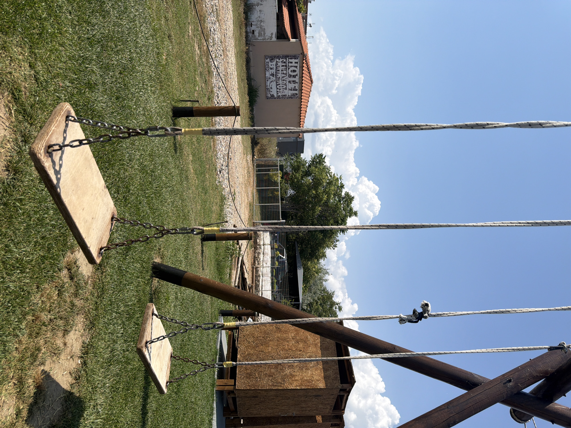 The image captures an outdoor scene on a bright, sunny day with a clear blue sky and fluffy white clouds, indicating midday or early afternoon due to strong shadows. In the foreground, two wooden swings hang from a rustic wooden swing set, attached by metal chains and ropes. The swings are empty, facing towards a grassy area and a gravel path.
In the mid-ground, a small wooden shed-like structure is visible, partially obscured by the swing set. Beyond this, a light-colored building with a terracotta tiled roof stands against the skyline. The building's side features a prominent mural composed of a grid of stylized letters and symbols. The word "HOTEL" is clearly visible in English on the upper right side of this mural. Other discernible elements within the grid include partial English words like "SANTA" and various Greek letters such as Lambda (Λ), Psi (Ψ), Theta (Θ), and Omega (Ω), suggesting a blend of languages, possibly for decorative or artistic effect.
Green grass covers much of the ground, interspersed with areas of dry earth and gravel paths. A tall tree with dense green foliage is visible to the right of the building, and a fence runs along the perimeter of the area. No people are present, depicting a quiet, recreational space in Alexandroupolis, Greece.