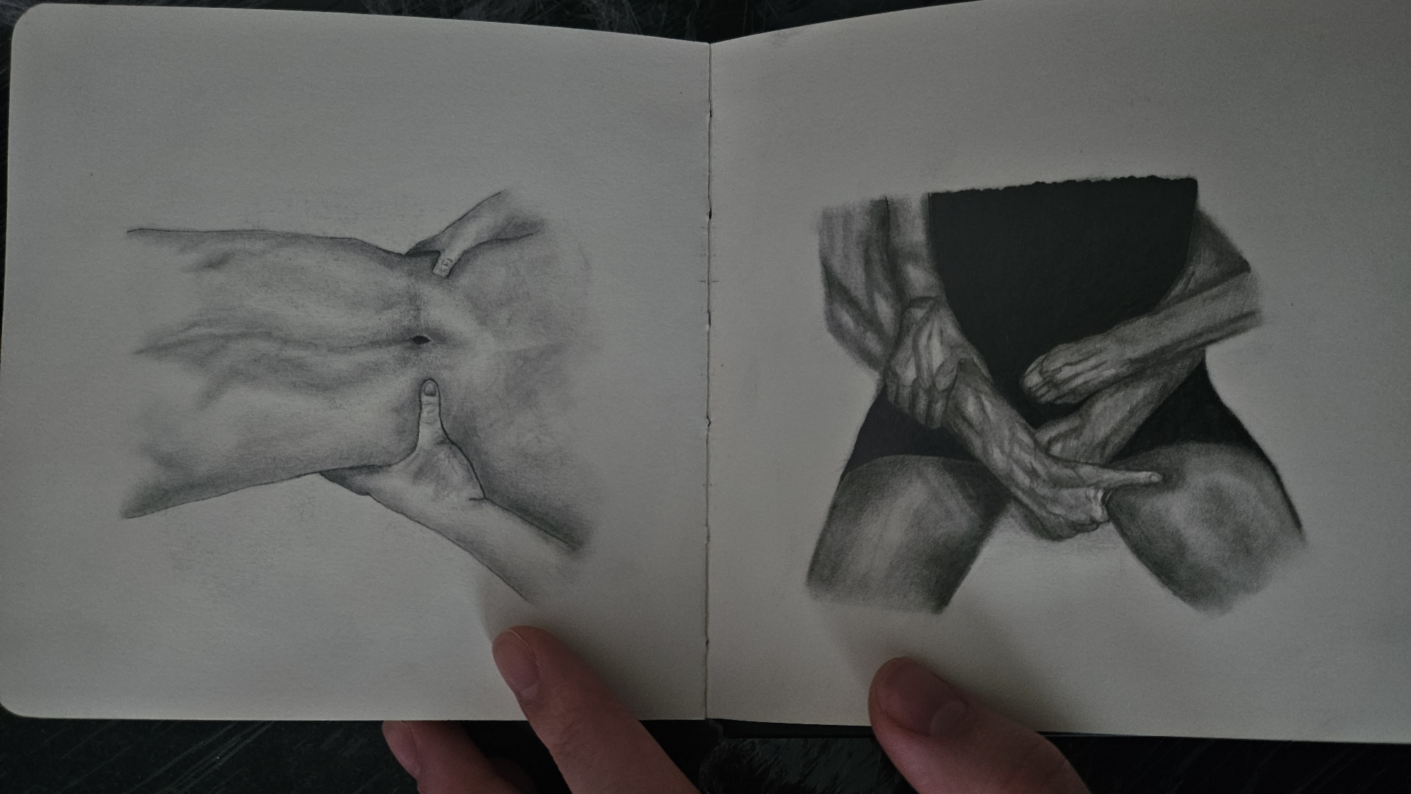 The image shows two pencil sketches in an open sketchbook. The top sketch depicts the lower torso and back of a person, with hands on either side of their waist. The bottom sketch shows a close-up of intertwined limbs, possibly arms and legs, with detailed rendering of muscles and skin texture. Both sketches are in grayscale and are highly realistic. The sketchbook is lying on a dark surface, and parts of a person's hand are visible holding the sketchbook open. There is no indication of a specific location within a city.