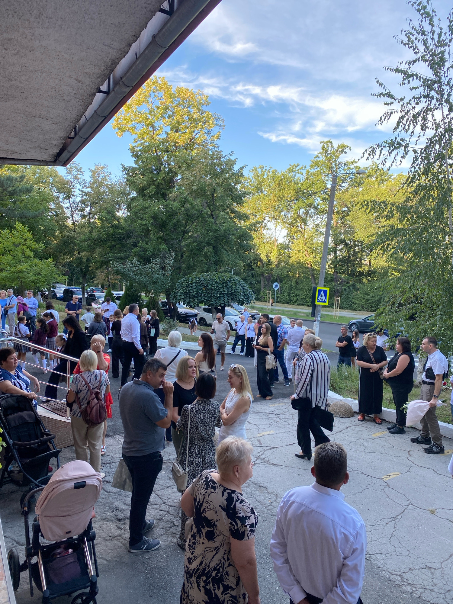 A large group of people are gathered on a concrete walkway outside a building with a covered overhang, surrounded by trees and parked cars. The scene appears to be in Chișinău, Moldova, indicated by a traffic sign with the number "30" and a pedestrian crossing symbol. Most individuals are dressed casually, with some in formal attire, suggesting a social gathering or an event. People are standing and conversing in small groups. There are two strollers visible in the foreground. The lighting suggests it is daytime, possibly late afternoon, with sunlight filtering through the trees.