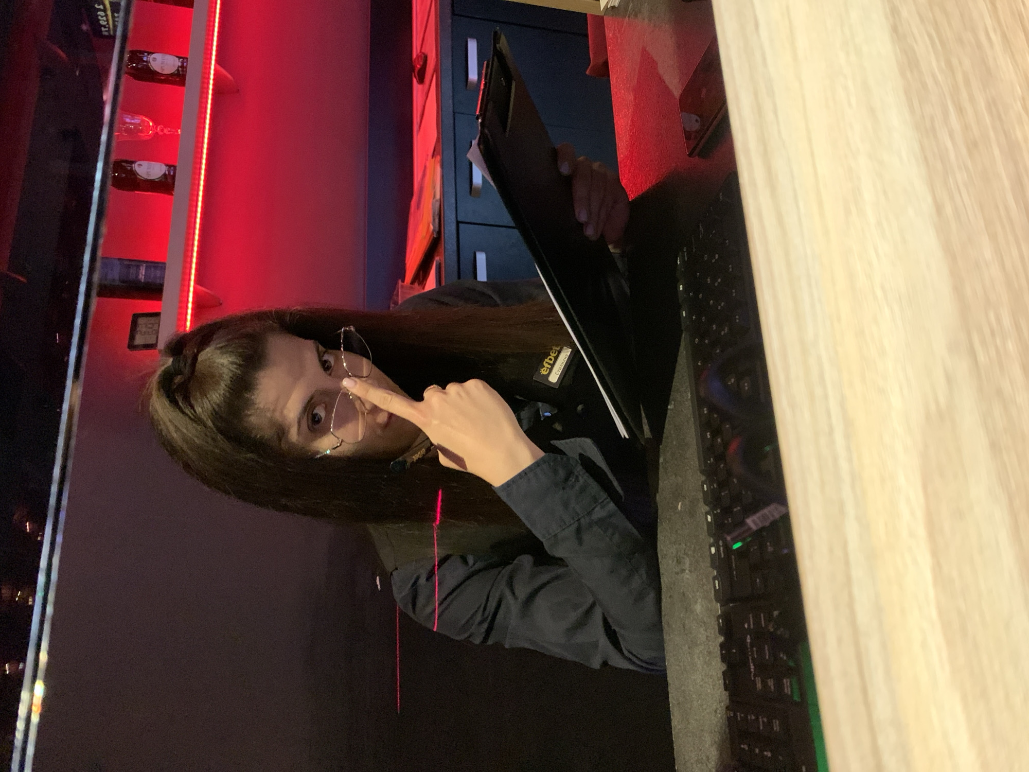 A woman with dark hair pulled into a high ponytail and wearing glasses and a dark shirt with a vest is leaning forward behind a desk. She is touching her nose with her right index finger. She is holding a black clipboard in her left hand. A keyboard with green backlighting is visible on the desk in front of her. Behind her, shelves are illuminated with red lighting, displaying bottles and glasses. The scene appears to be an indoor establishment, possibly a bar or club. The context provided suggests the location is in Burgas, Bulgaria.