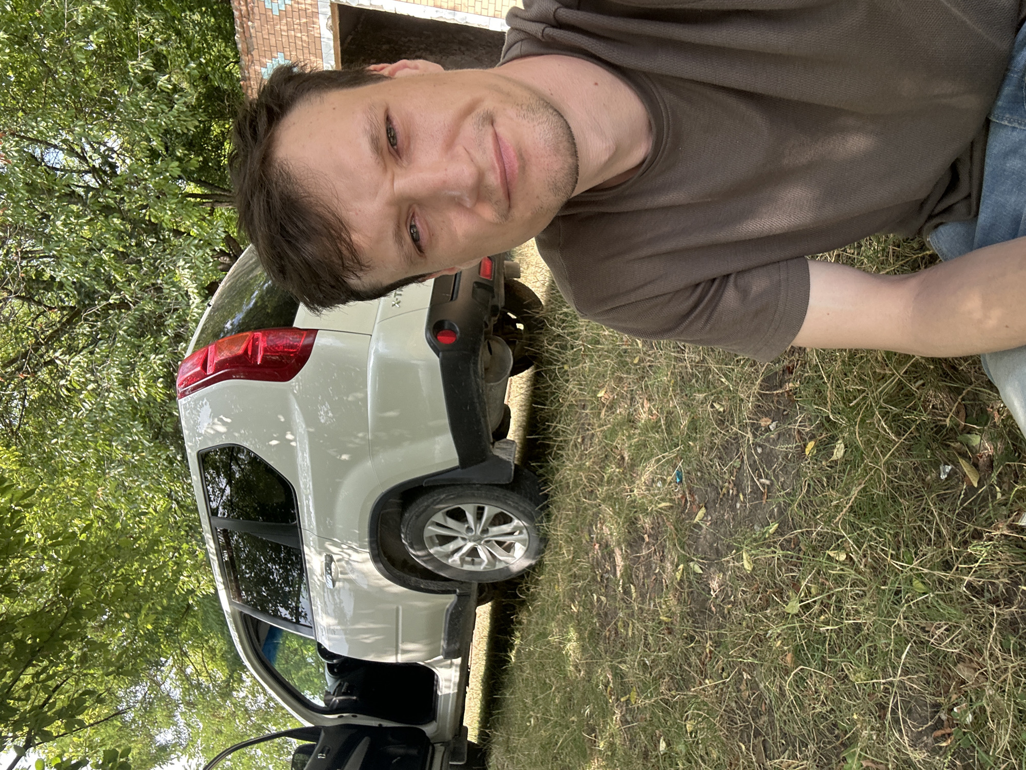 A man in a brown t-shirt and blue jeans sits on the ground in front of a white SUV parked outdoors. The SUV's driver-side door is open, revealing the interior. The vehicle is positioned in front of a brick structure and surrounded by trees and dry grass. The scene is likely located in Makiv, Ukraine, given the context. The man is looking directly at the camera, with a slight smile.