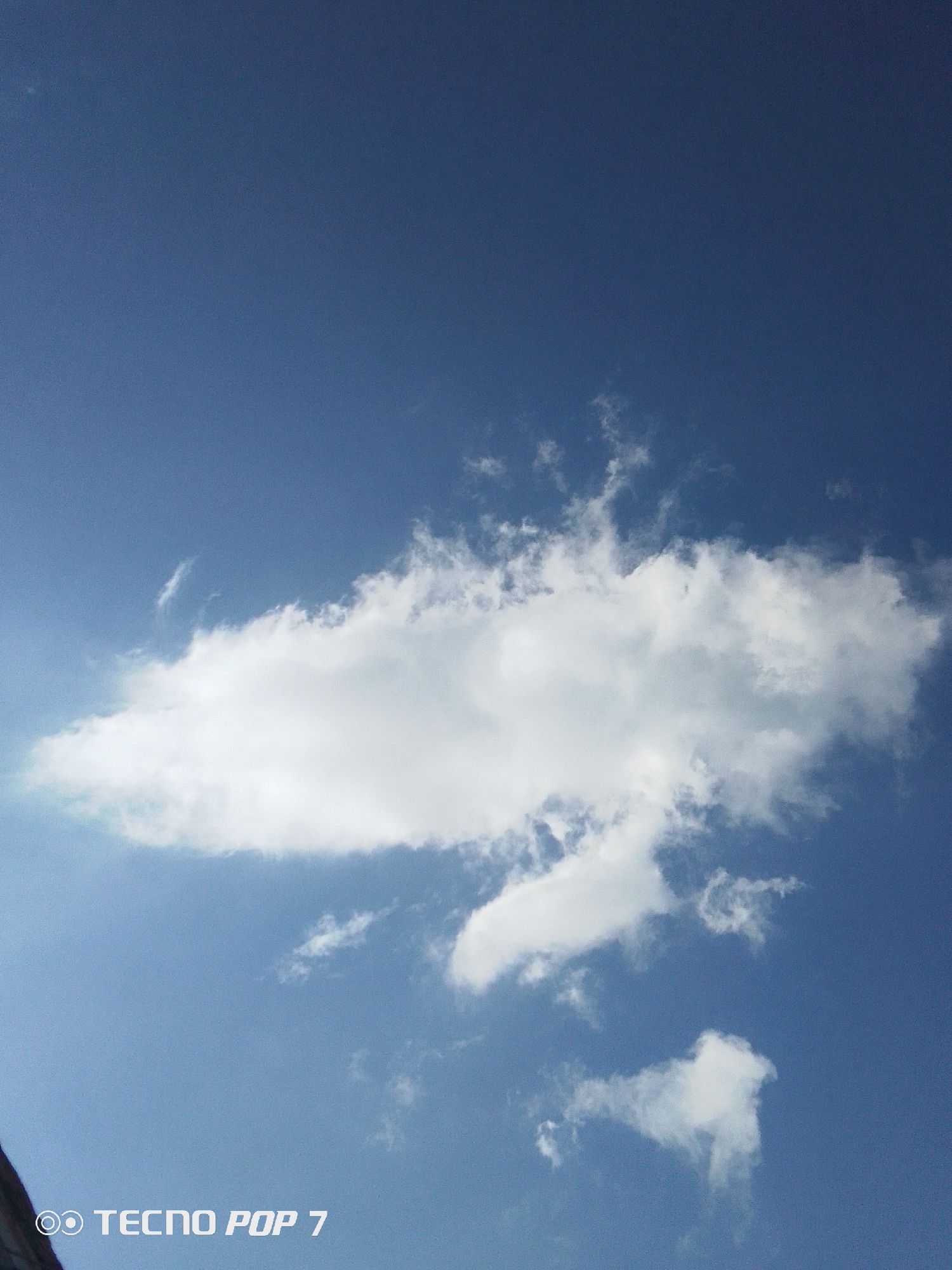 The image shows a blue sky with white clouds. A large cloud dominates the center of the image, with smaller clouds below it. The bottom left corner of the image shows part of an unknown object and the watermark "TECNO POP 7" which suggests the image was taken with a mobile phone. There are no indications about the city of Pedro González, Venezuela.
