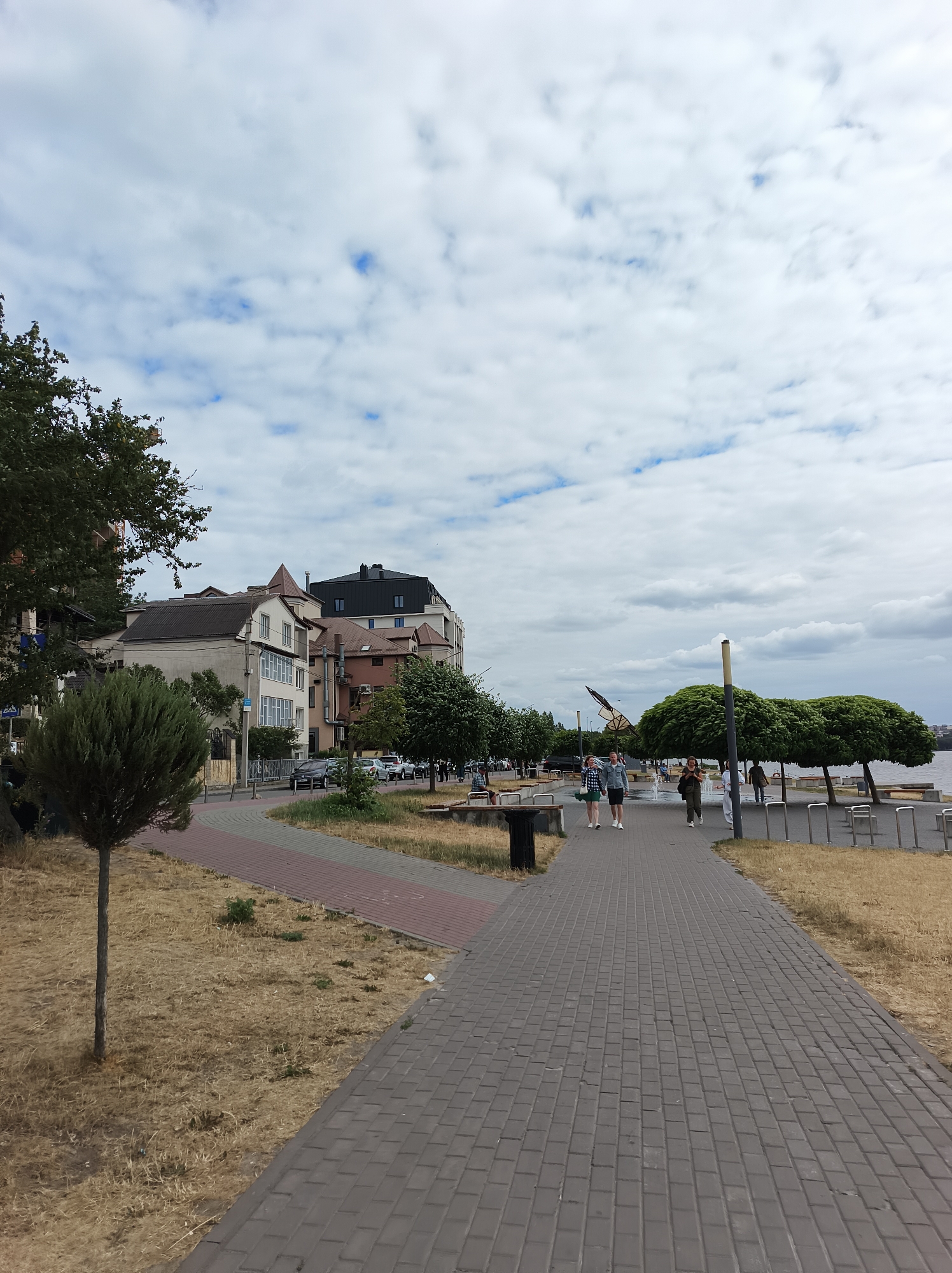 This image depicts a view of Ternopil, Ukraine, focusing on the embankment of Ternopil Lake. A paved walkway, constructed from grey interlocking bricks, dominates the foreground and extends into the distance. To the left of the walkway, dry, light-brown grass covers the ground, sparsely dotted with small green plants and a single slender tree with a conical canopy. Adjacent to the grass is a curved path made of red and grey rectangular pavers. Beyond this path, several multi-story buildings with varying architectural styles and rooflines are visible, interspersed with greenery. Cars are parked along the street in front of the buildings.
To the right of the main walkway, a body of water, presumably Ternopil Lake, is visible. Several people are walking along the embankment. Some are in small groups, while others walk individually. The people are casually dressed. The embankment is lined with benches and lampposts. Several trees with rounded, dense canopies are planted along the water's edge. The sky is overcast with a mixture of white and grey clouds, with patches of blue visible.