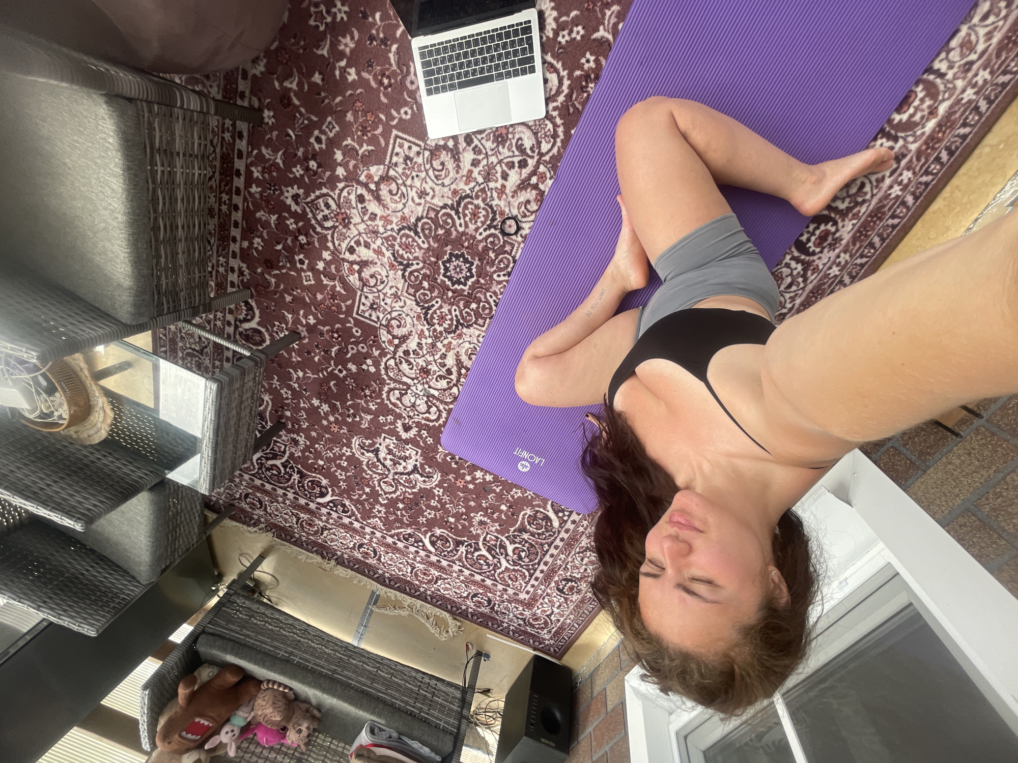 A woman is lying on a purple yoga mat on an ornate rug. A laptop is on the rug next to the mat. There is a wicker chair and table set with stuffed animals on the left of the scene and a speaker near the bottom of the rug and near a brick wall with a window. The scene appears to be on a balcony or patio. The city of Мосметаллоконструкция Онт, Russia is not discernible from the image.