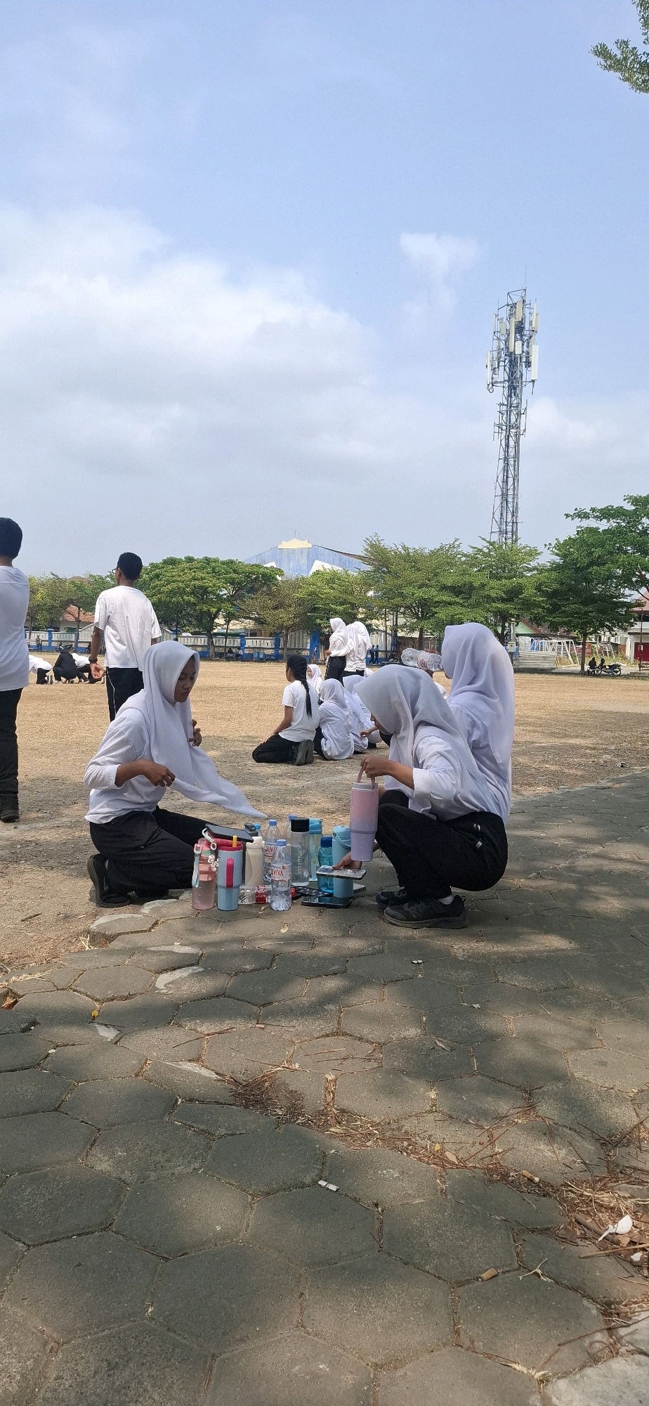 In a wide, outdoor field, likely a sports ground, several individuals, predominantly young women dressed in white shirts and hijabs, are gathered. They are seated on the dry, brown earth, some in a crouched position. In the foreground, two women are interacting with a collection of items laid out on the ground. These items include several plastic water bottles, a pink and blue tumbler, and what appears to be some sort of container or box with blue lids. One woman is holding a pink tumbler, while the other is holding a white scarf. The ground they are sitting on is paved with hexagonal concrete blocks in the immediate foreground, transitioning to a dusty, sandy soil further back.
In the middle ground, other individuals are also seated or squatting, some facing away from the camera. Behind them, a line of trees with green foliage separates the field from buildings in the background. One building has a distinctive blue and white facade. Dominating the sky in the upper right quadrant of the image is a tall, multi-tiered telecommunications tower. The sky above is a pale blue with scattered white clouds, suggesting a bright, sunny day. The overall environment appears to be a public outdoor space, potentially a school or training ground, located in Baturan, Indonesia, as indicated by the distinct vegetation and architectural style.