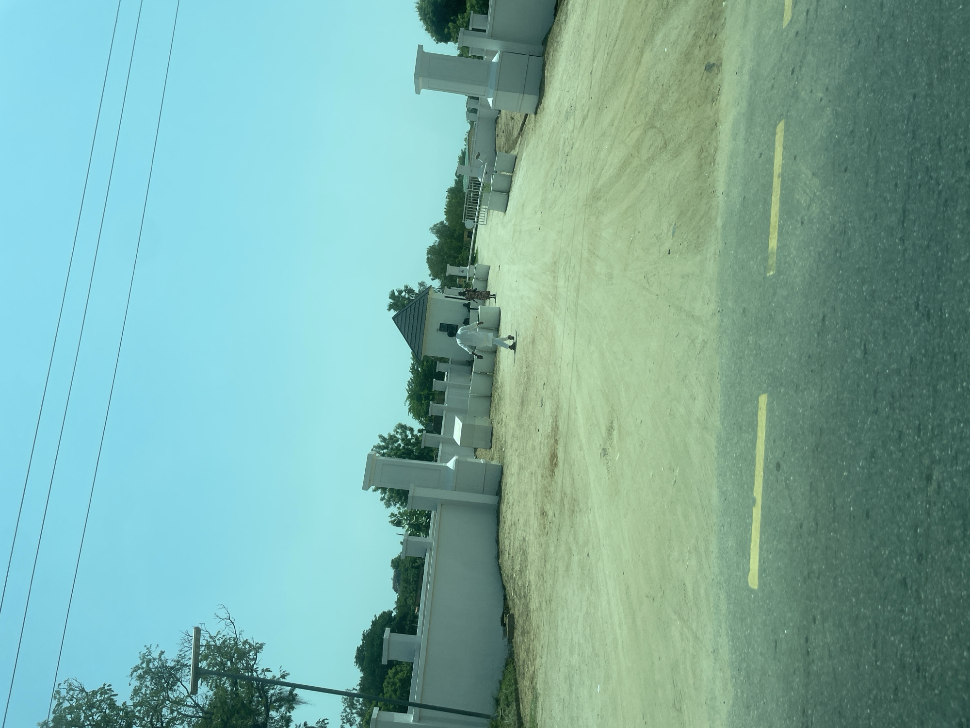 The image depicts an outdoor scene with a road on the right side, marked with yellow lines. The road is adjacent to a dirt path. On the left side of the path, there are several white structures, some of which appear to be modern buildings with angular designs and decorative elements. Trees and foliage are visible behind these structures, suggesting a rural or semi-urban setting. The sky is clear and bright, indicating a sunny day.
Two people are visible on the dirt path. One person, dressed in white, is walking away from the camera. Another person, wearing patterned clothing, is also walking. The presence of power lines in the upper left corner of the image provides a common visual cue. The overall impression is a snapshot of daily life in a location undergoing construction or development, possibly a residential area or a commercial space with modern architectural elements. The time of day appears to be daytime, given the strong sunlight and clear sky. The location context of Bauchi, Nigeria, suggests this scene is set in that region. No text is visible in the image.