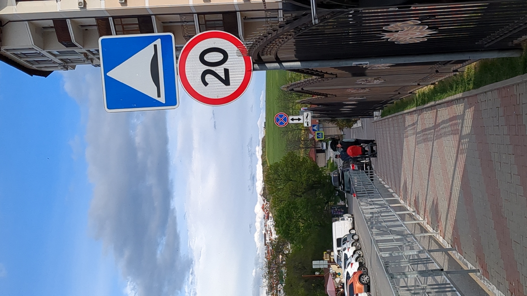 The image shows a street scene in what is possibly the city of Gelendzhik, Russia, based on the Cyrillic signage. There are two street signs on a post: one indicating a speed bump ahead (blue square with white symbol) and another indicating a speed limit of 20 (presumably kilometers per hour). A wrought-iron fence runs along the right side of the frame, separating the sidewalk from a grassy area. A paved sidewalk stretches along the right edge of the frame. Beyond the sidewalk, cars are parked along the curb. Several individuals are walking on the sidewalk. In the background, there is a field, some buildings, and trees under a sky with clouds. The image appears to be taken from an elevated position.