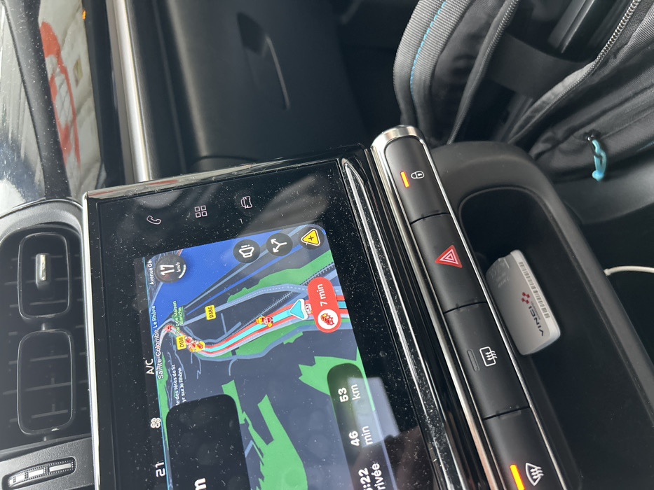 The image displays the interior of a vehicle, focusing on the central infotainment screen and a portion of the dashboard. The navigation system on the screen indicates the vehicle's current location on the D138 road in Saint-Cyr-sur-le-Rhône, France, with nearby textual references to Sainte-Colombe and Le Rhône, as well as Parcelle Maison Blanche and D386. The vehicle is moving at 17 km/h, with an estimated 46 minutes and 53 km remaining to the destination; a 7-minute delay is also indicated.
Below the screen, a row of control buttons is visible. The rear defroster button and the central door lock button are illuminated orange, signifying their active state. Beneath these controls, a small open compartment contains a white "VINCI Autoroutes" electronic toll transponder with a barcode visible on its side. Two horizontal air vents are positioned above the infotainment screen. A black bag, featuring blue stitching and a light blue zipper pull, is partially visible in the lower right corner of the frame. No individuals are depicted in the scene.