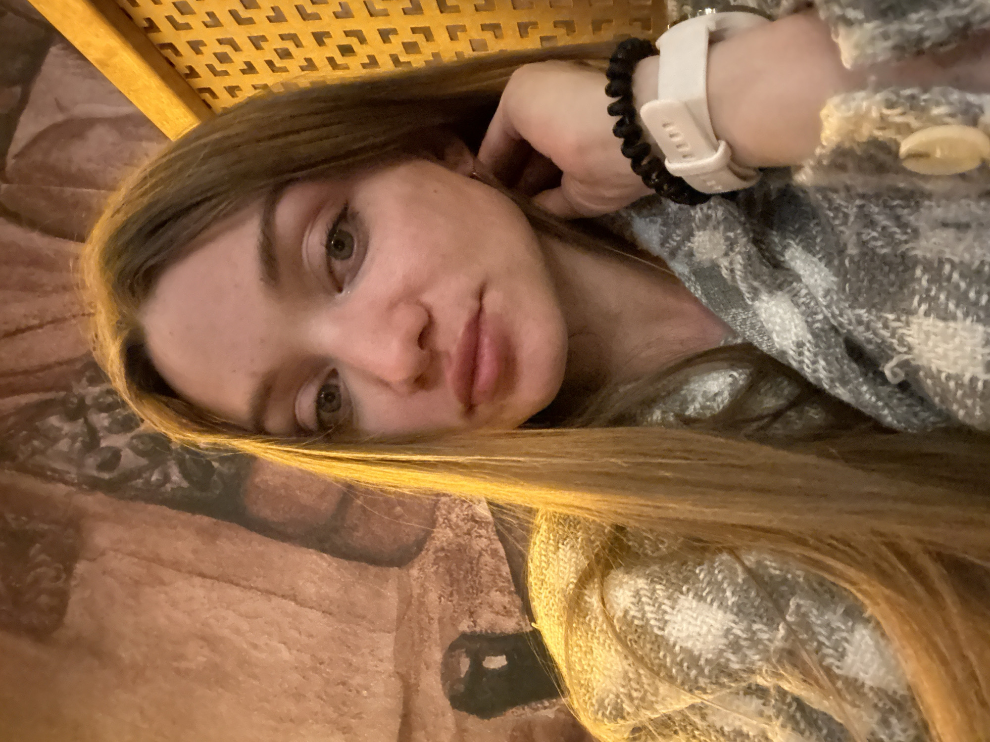 A selfie of a young woman with long blonde hair, wearing a gray plaid sweater. She has a white smartwatch and a black beaded bracelet on her left wrist. The background includes a wall with indistinct patterns and a woven, lattice-like structure above. The location is identified as Kazan, Russia.