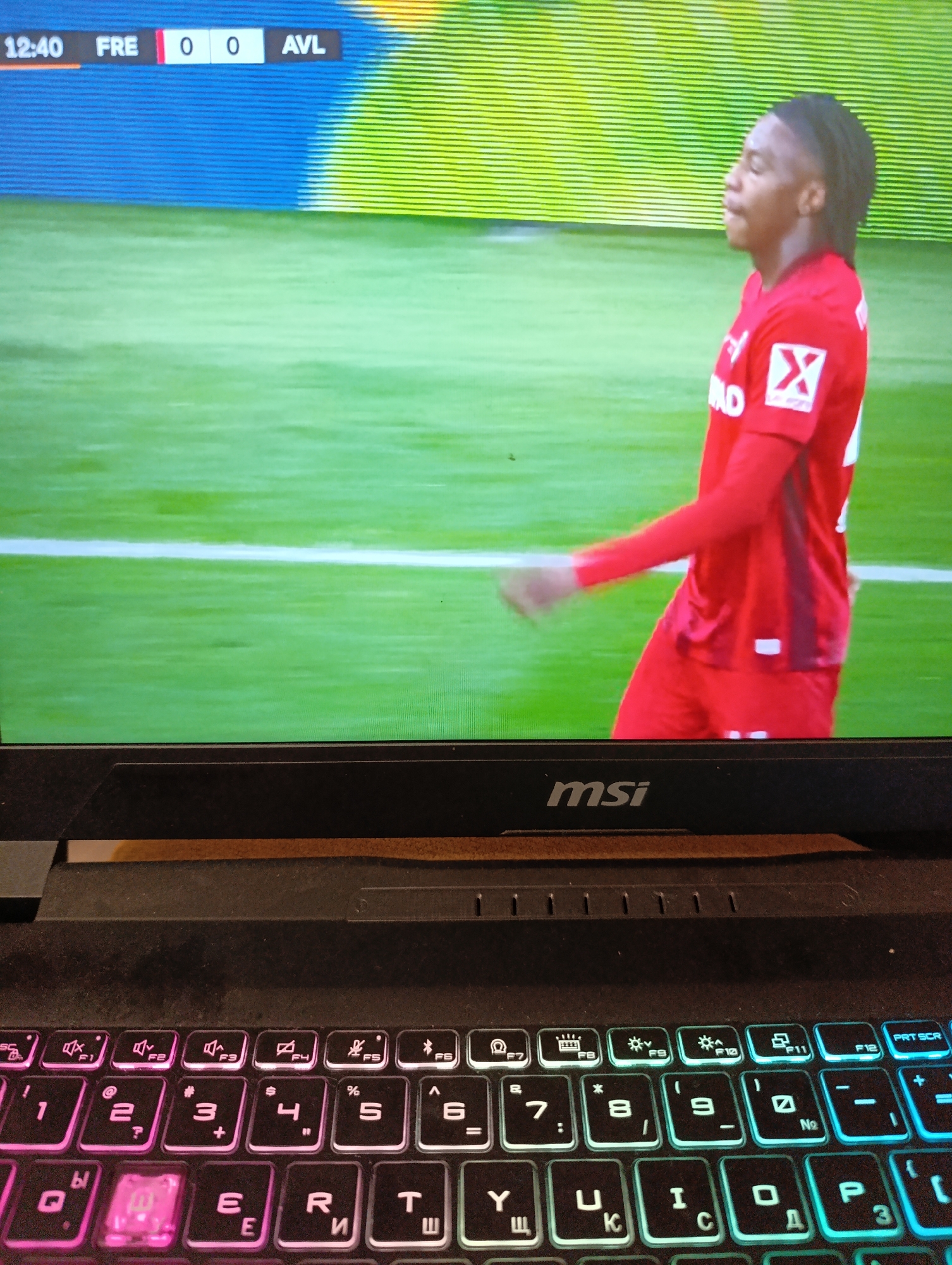 The image shows a laptop with an MSI brand name visible below the screen. The laptop keyboard is lit with a rainbow effect, featuring pink, white, and blue sections. On the screen, a soccer game is visible. The score is tied 0-0 between FRE and AVL, at 12:40. A soccer player in a red jersey is standing on the field. The environment appears to be a broadcast or recording of a soccer match. The image does not show any location details for Plovdiv, Bulgaria.