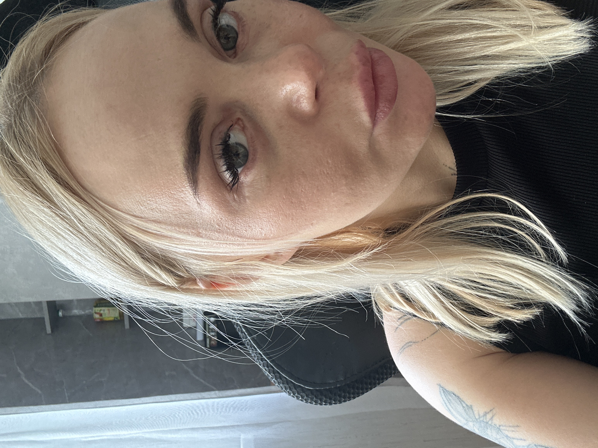 The image is a close-up indoor portrait of a fair-skinned female with long, straight, blonde hair. Her eyes, appearing light green or hazel, gaze directly forward, complemented by dark, defined eyebrows and prominent, dark eyelashes. Her lips are full and tinted a muted pink, slightly parted. Visible skin texture includes fine lines on the forehead and pores on the cheeks. She wears a black, ribbed-knit top. A faded blue-grey tattoo featuring a feathered design is visible on her left upper arm, and a small, faint mark, possibly a tattoo, is present on her neck below her left jawline.
The background reveals a domestic or office setting. To the left, a sheer white curtain covers a window, admitting soft, diffused natural light. Behind her left shoulder, a dark-colored office chair with a textured backrest is partially visible. The wall behind the subject is light grey with a subtle dark grey marbled pattern. Two minimalist shelves are mounted on this wall: the upper shelf holds a clear glass bottle with a light yellow liquid and an indistinguishable label, while the lower shelf contains indistinct small objects.