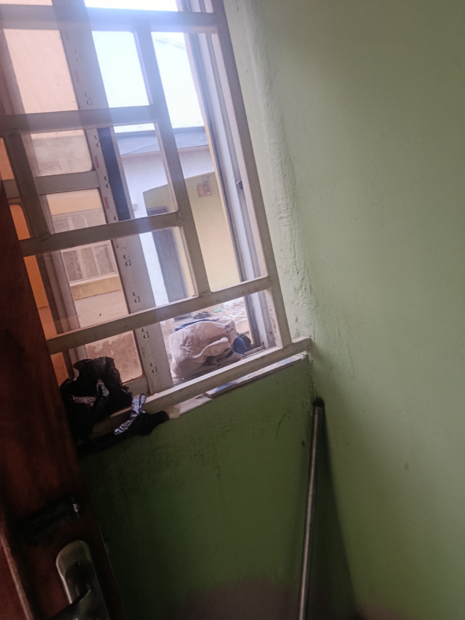 The image shows an interior scene looking out a barred window. The window has metal bars that divide the view into several rectangular panes. Through the window, other buildings and structures are visible, suggesting an urban or semi-urban environment. In the foreground, a portion of a green wall is visible, and to the left, a dark brown door frame with a metal handle is present. On the windowsill, there are some crumpled items, including what appears to be a black plastic bag and some light-colored fabric. The lighting suggests it is daytime, with sunlight streaming through the window, creating bright highlights and some glare. The overall impression is of a somewhat cluttered and utilitarian space, viewed from an interior.