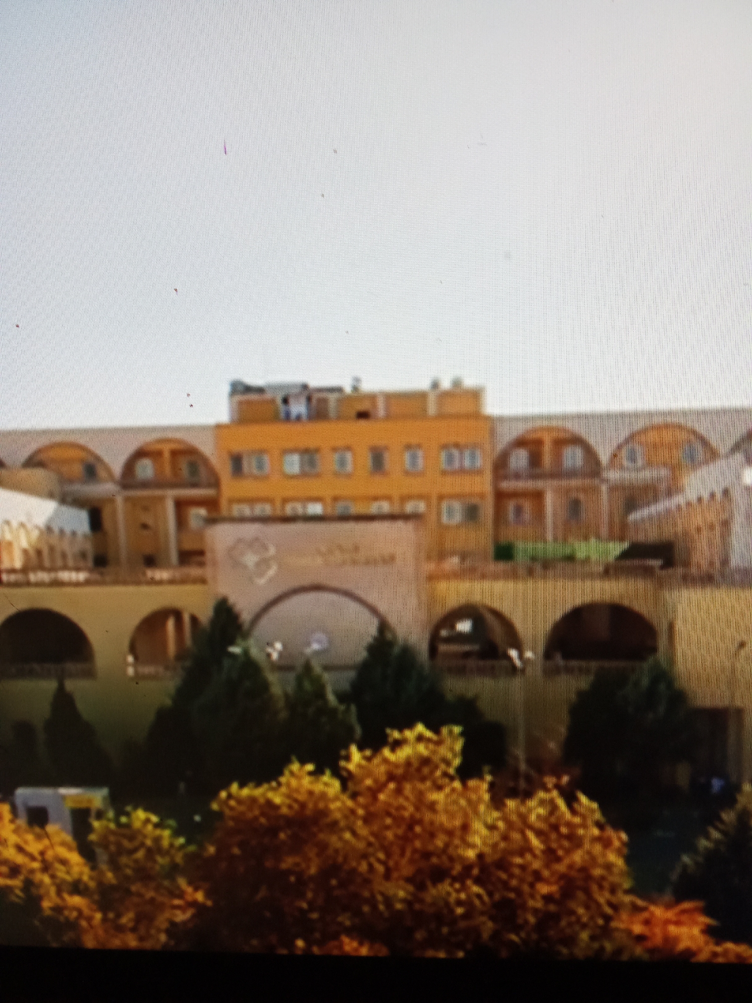 This is a blurry, medium-shot, eye-level photograph of a building complex, possibly a hotel or a residential building, with a clear sky. The architecture is characterized by arched windows and a warm, yellowish-orange facade. In the foreground, there are trees with bright yellow and orange foliage, suggesting an autumn setting. A white vehicle with yellow markings is partially visible to the left of the frame. The building has multiple levels and appears to be designed with a sense of grandeur, featuring recessed sections and decorative elements. The overall impression is of a well-maintained, possibly resort-like property. The location is identified as Рощин ауылдық округі, Kazakhstan.