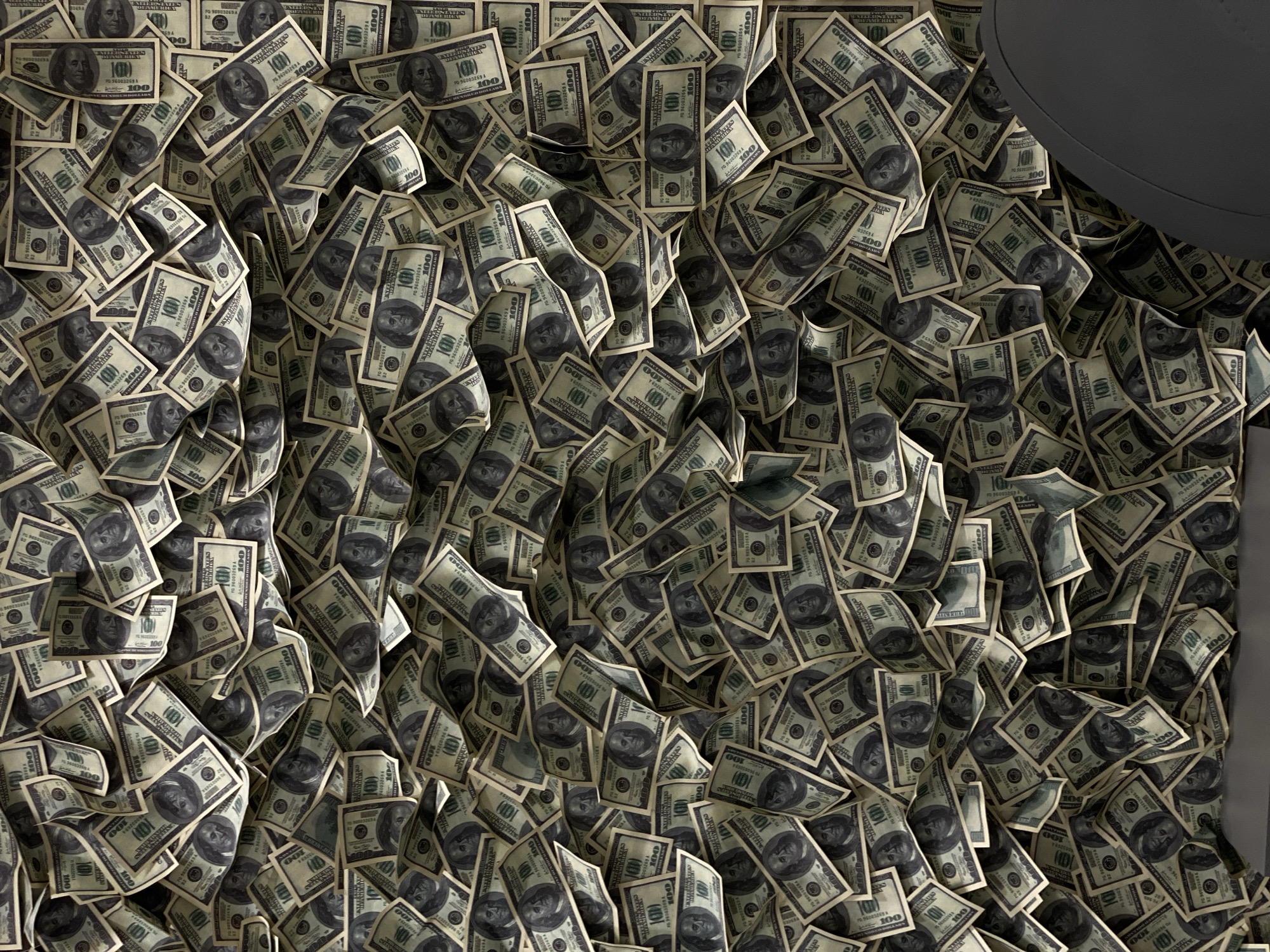 The image depicts a large quantity of $100 US dollar bills scattered on a surface, with a portion of a gray, rounded object visible in the upper right corner. The bills are arranged haphazardly, overlapping and facing different directions. No people or other subjects are apparent. The scene appears to be staged and lacks any identifying features to confirm a location, including Sochi, Russia.