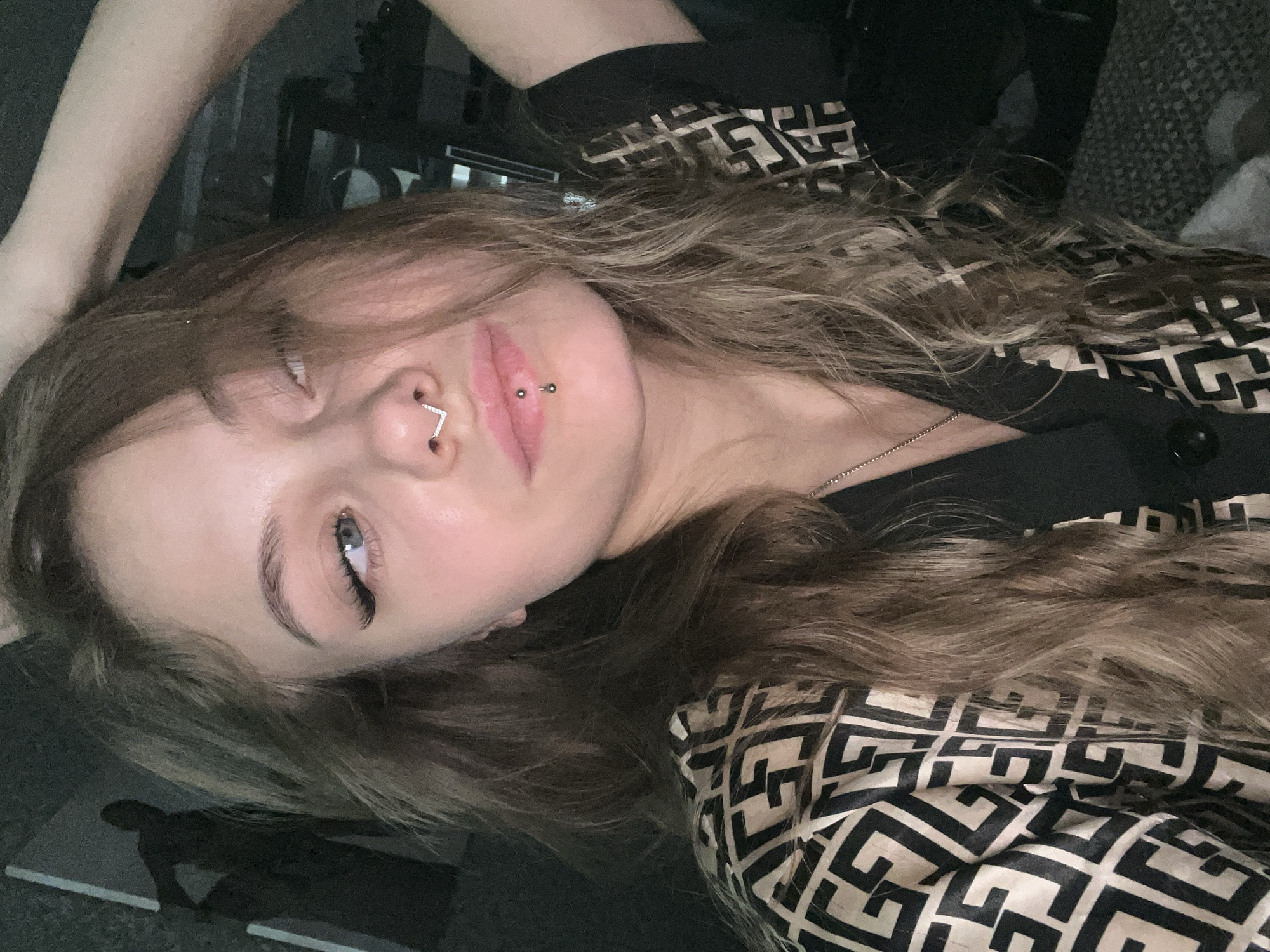 A young woman with light brown, wavy hair and pale skin is lying down, looking slightly upwards with her left eye open. She has a piercing in her nostril shaped like a right angle and two piercings in her lower lip. She is wearing a black and tan patterned top. The background is dark and out of focus. The image doesn't suggest any specific information that confirms the city of Фоки, Russia.