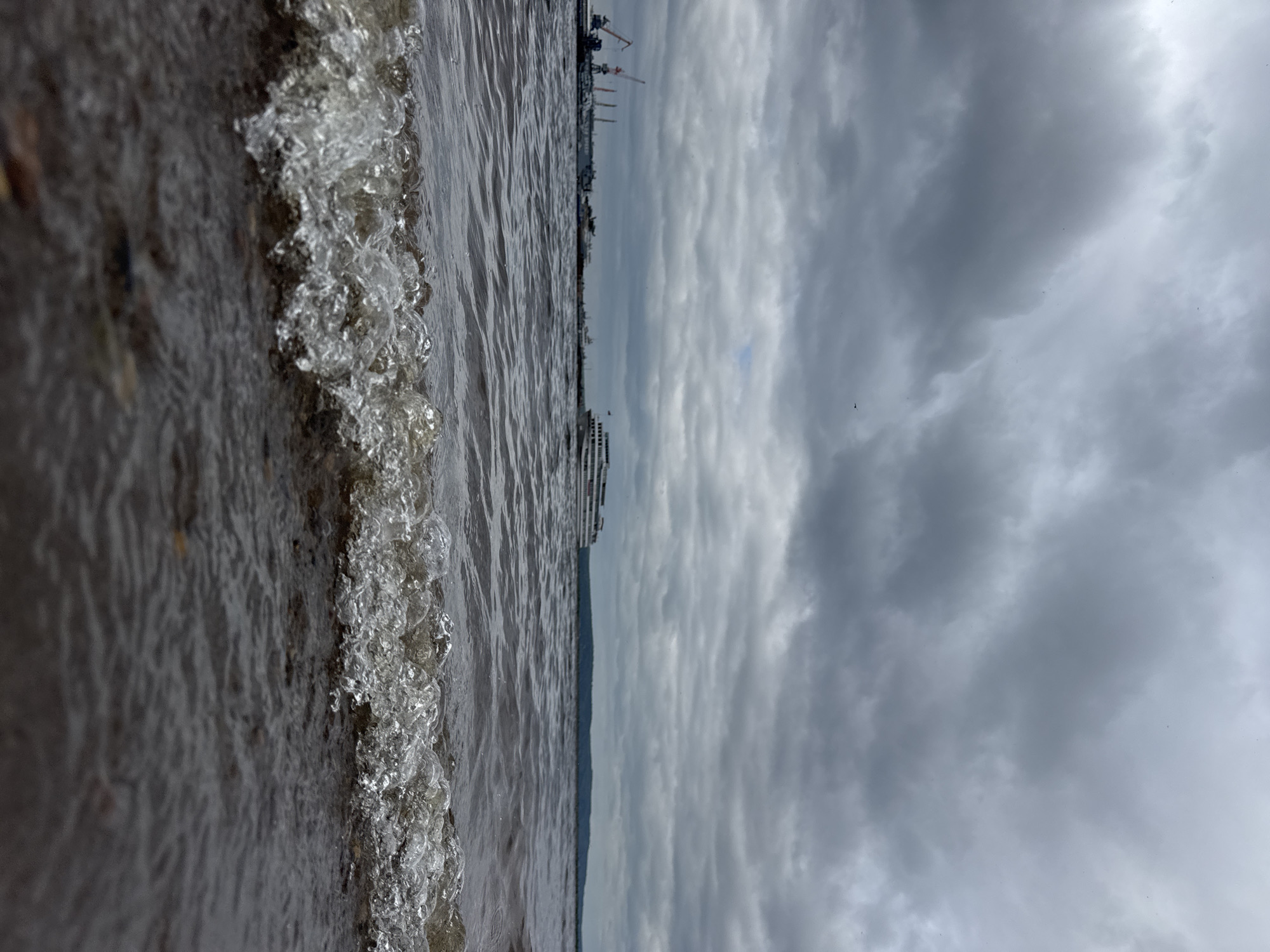 The image depicts a cloudy day at the Kama River in Chaikovsky, Russia. In the foreground, a small wave with visible bubbles is breaking on the pebbly shore. In the middle ground, a large white passenger ship is sailing on the water, its decks and windows clearly visible. Further in the background, a port is visible with several cranes and other vessels docked along the shore. The sky is overcast with thick, gray clouds, and the distant horizon is marked by a strip of land. The overall scene conveys a sense of a calm, overcast day by the river.