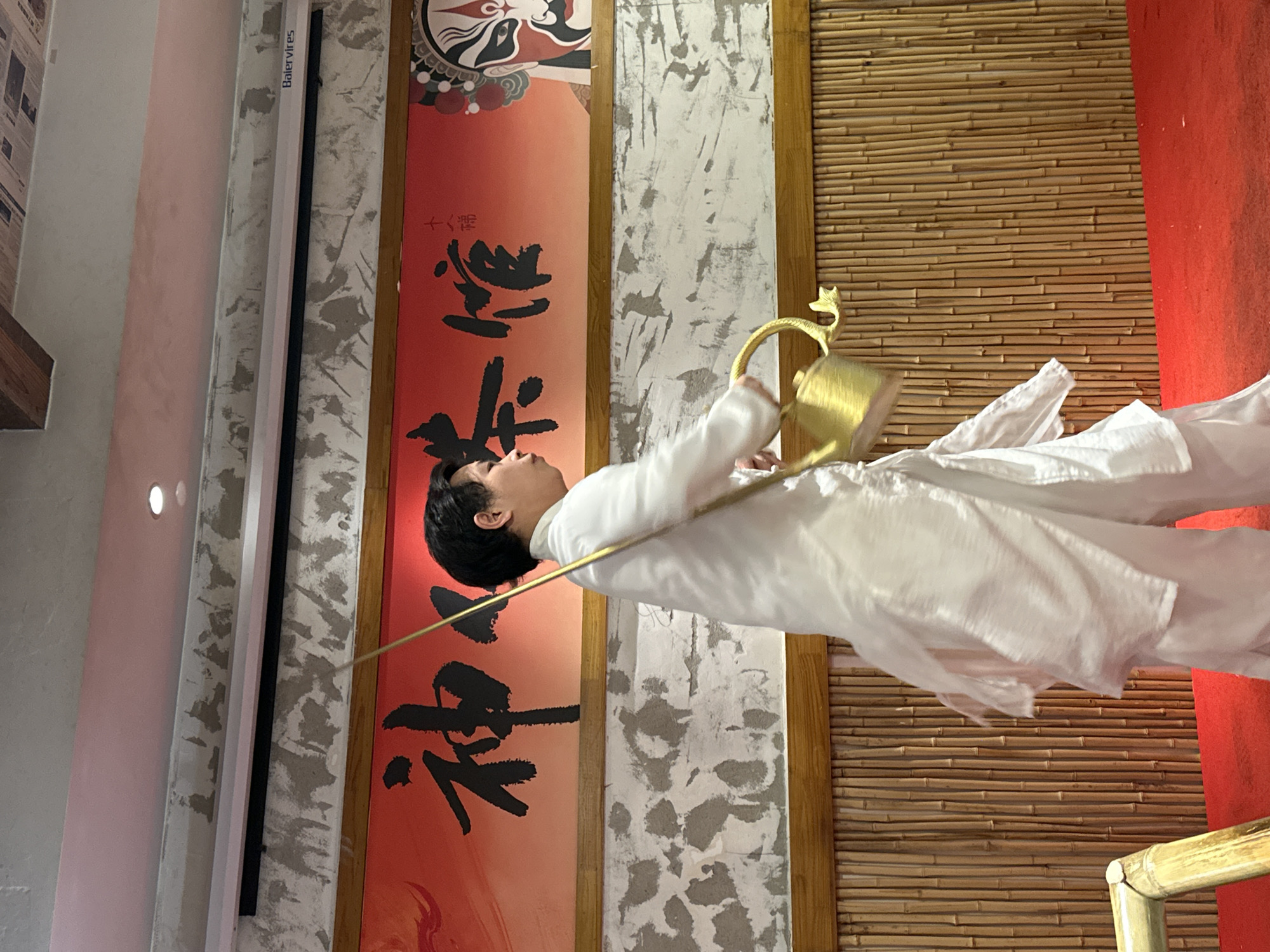 A person in white clothing is holding a golden teapot with a decorative handle in front of a bamboo wall. A red banner with Chinese calligraphy is displayed on the wall beside the bamboo, next to a gray and white wall. The scene is located indoors. Based on the writing, this could be in China. More specifically, the image may have originated in Yuzhong District, China.
