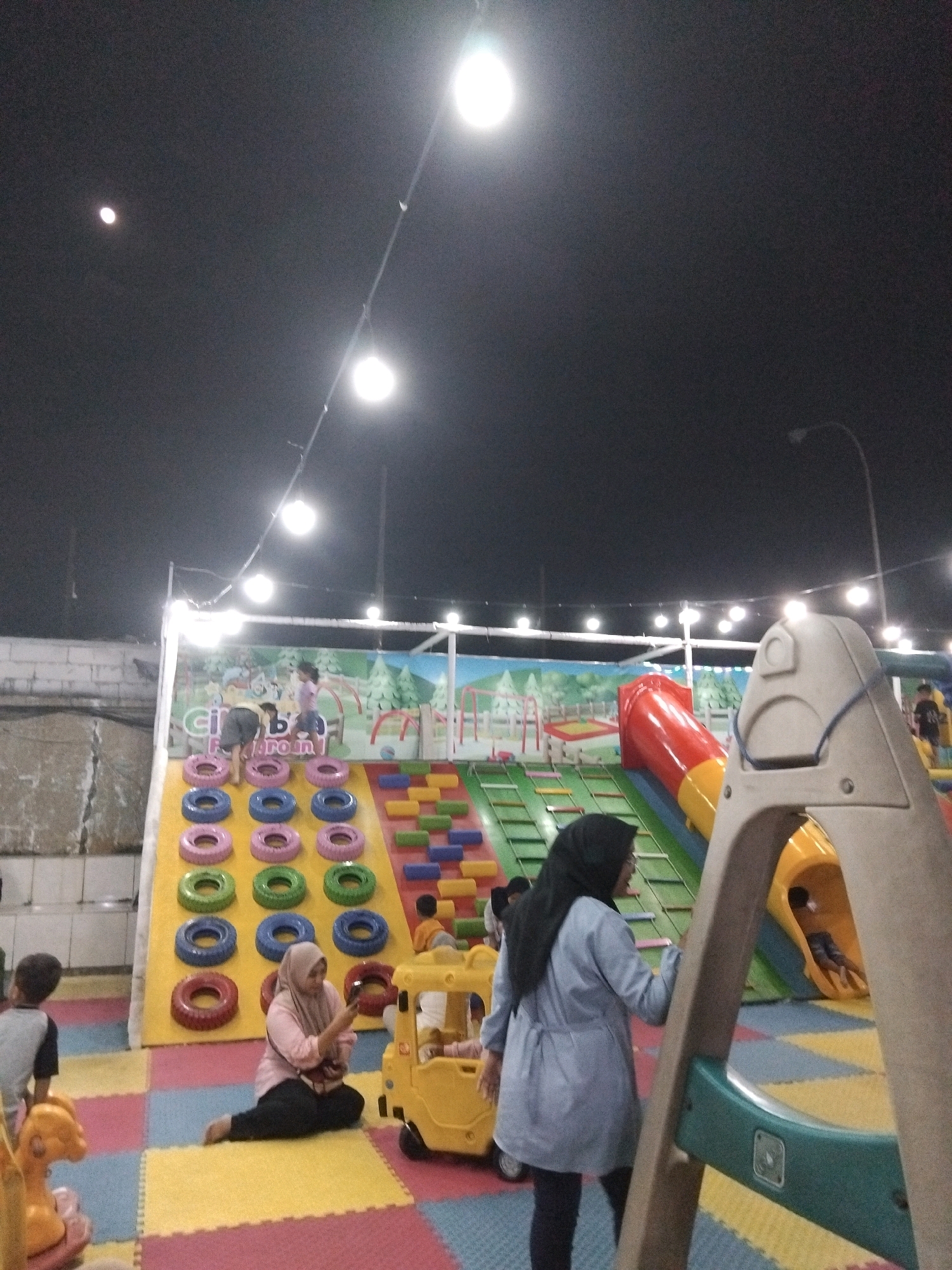 The image depicts an indoor playground at a location in Sedati Agung, Indonesia. The scene is illuminated by overhead lights. In the foreground, a woman wearing a black hijab and a light blue jacket is standing. To her left, another woman in a light pink hijab and a pink shirt is sitting on the floor, looking at her phone. A yellow toy car is next to her. Behind them is a large, colorful play structure with slides, climbing walls made of colorful rings and blocks, and a red tube slide. Several children are visible playing on the structure. In the background, a large mural with a cartoon landscape is painted on the wall. The floor is covered with a mat of interlocking colorful squares.